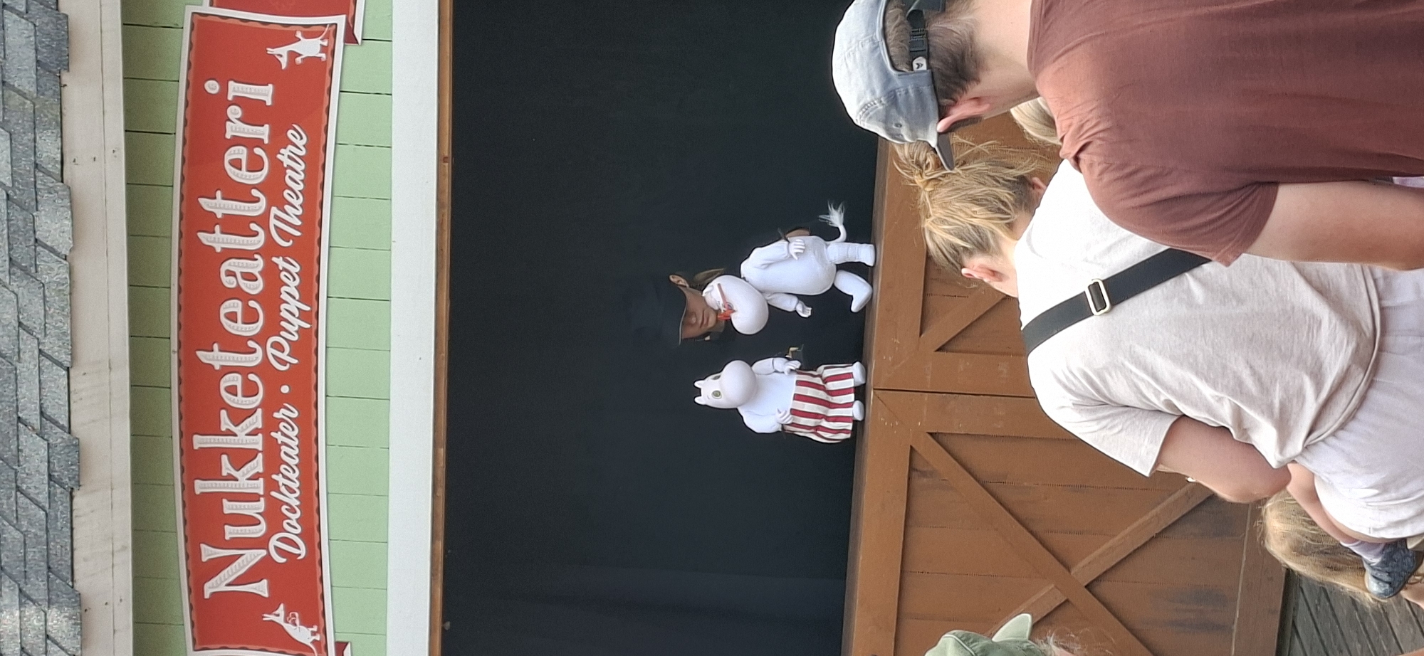 The image shows a puppet theater stage at Moomin World in Naantali, Finland. Two Moomin puppets, one wearing a red and white striped skirt and the other in white, are visible on stage, seemingly being operated by a person partially obscured behind them. The stage has a wooden facade. In the foreground, several people are watching the puppet show. The sign above the stage reads "Nukkeateatteri Dockteater · Puppet Theatre." The Moomin characters are from the Moomin stories created by Finnish author Tove Jansson.