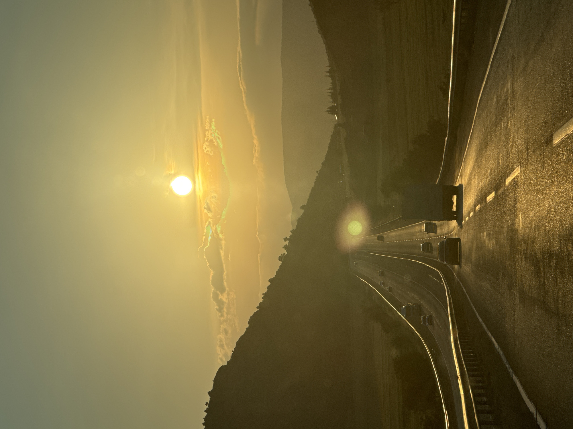 The image depicts a multi-lane highway traversing a mountainous landscape in Korkuteli, Turkey, under a sunset or sunrise. The prominent, intense golden sun, positioned low in the sky slightly above the horizon, casts a warm, backlighting glow across the entire scene, resulting in significant lens flare. The sky transitions from a bright yellow-orange near the horizon to a duller yellowish-green higher up, with scattered clouds partially silhouetted.
On the left, a dark, undulating mountain range slopes downward towards the road, covered sparsely with silhouetted trees. A similar but more distant, hazy mountain range is visible on the right. The highway extends from the foreground into the distance, appearing to vanish at the horizon, highly reflective with the golden light. White solid and dashed lane markings are visible, and metal guardrails flank both sides of the road in the foreground, also reflecting the light.
Multiple vehicles are present on the highway. In the immediate foreground on the right, a large white semi-truck is traveling away from the viewer, followed by a dark sedan. Several other cars and another truck are visible further down the road, also heading into the distance, appearing as dark silhouettes against the bright backdrop. No distinct human figures are individually identifiable.