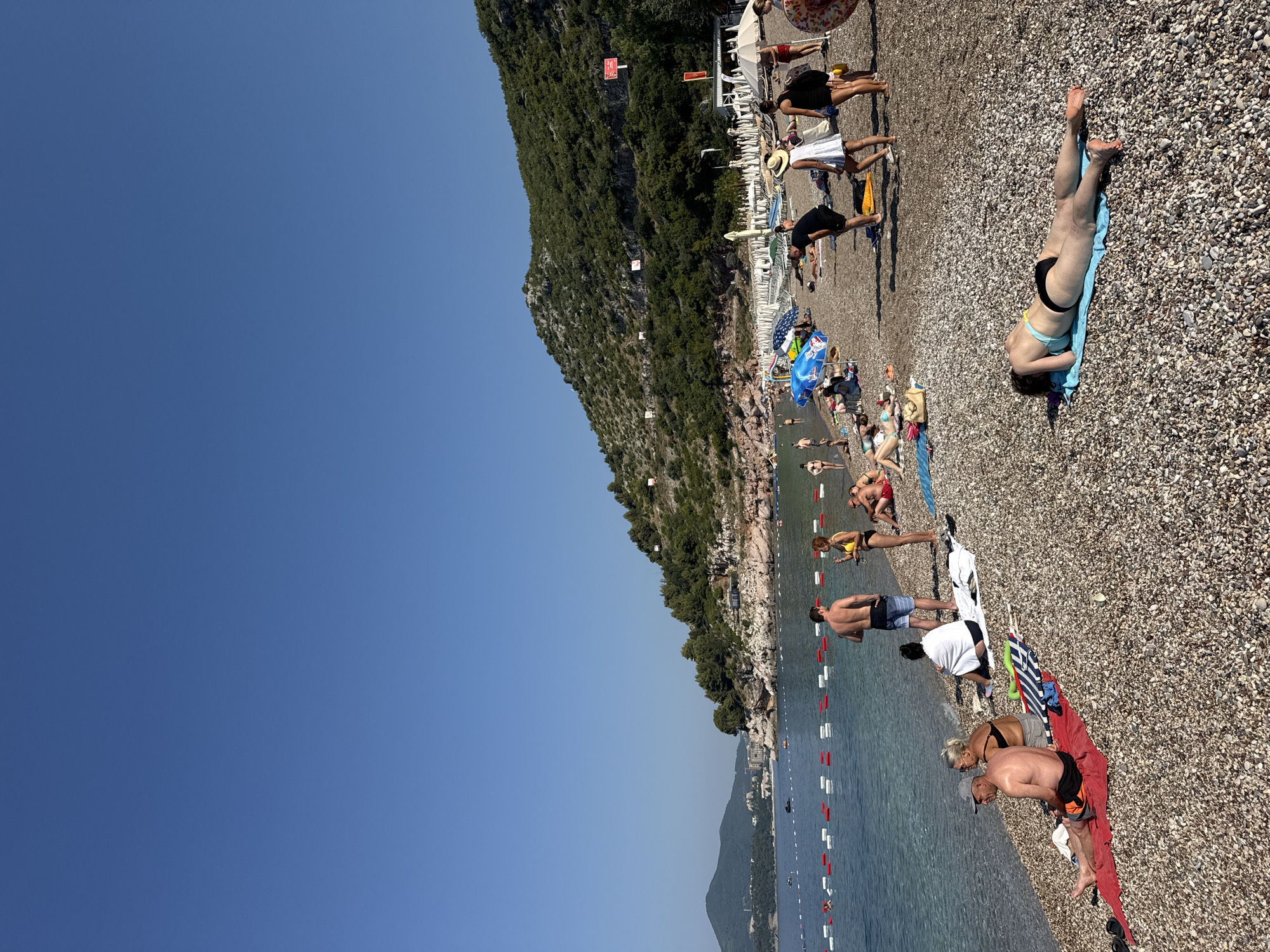This media file depicts a sunny day at Kamenovo beach in Montenegro. The scene is a crowded pebble beach with many people sunbathing and relaxing. The water is calm and clear, with a few boats visible in the distance. A steep, tree-covered hill rises behind the beach, and a small town or resort can be seen nestled along the coastline further down.
The people are dressed in swimwear, and some are lying on towels or sun loungers. A few individuals are partially in the water. The atmosphere appears relaxed and leisurely, typical of a summer beach day. The time of day is likely mid-afternoon, given the bright sunlight and the shadows cast by the people.
Visible objects include beach umbrellas, towels in various colors, and a demarcation line with buoys in the water. There are several white structures that appear to be part of a beach setup or facilities. Some red and white signs are visible on the hillside, but the text is too small to read.
The overall impression is one of a vibrant and popular tourist destination, enjoying warm weather and a beautiful coastal landscape.