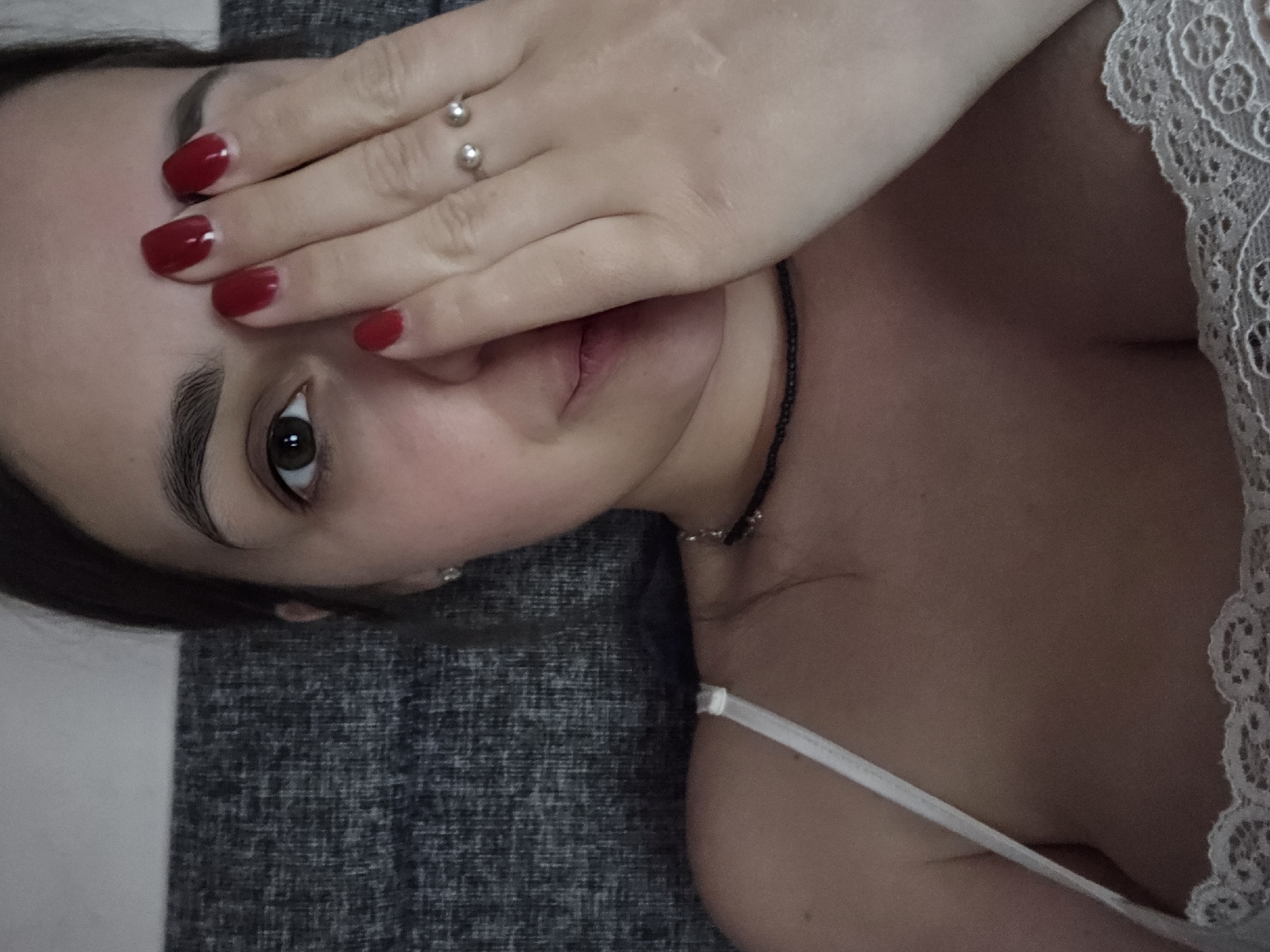 A close-up, waist-up selfie shows a woman with dark hair and brown eyes. She is wearing a white camisole with lace trim and a thin strap. Her right hand is covering her right eye, and her fingernails are painted red. She is also wearing a silver ring with two small pearls on her left hand. A thin black choker necklace is visible around her neck. The background appears to be a textured gray fabric, possibly upholstery. The lighting is dim, suggesting an indoor setting. There is no information to suggest the image was taken in Petropavlivska Borshchahivka, Ukraine.