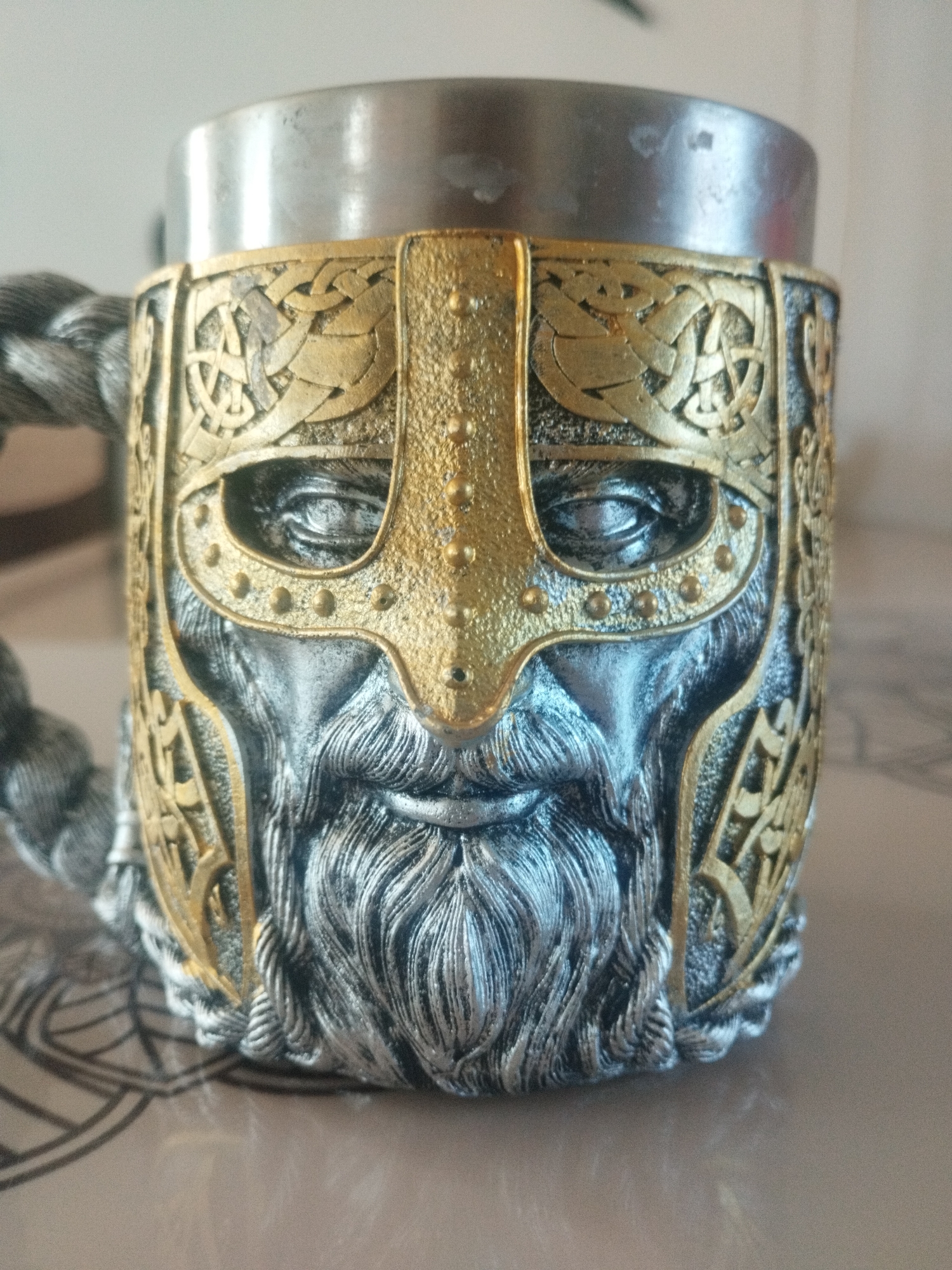 A close-up, eye-level shot shows the front of a metal mug with a detailed, sculpted design. The mug is made of silver-colored metal with gold accents. The design features a warrior's face with a helmet. The helmet is gold and has intricate knotwork patterns and is studded with small gold dots. The face beneath the helmet is silver and sculpted to show the eyes, nose, and a long, braided beard. The eyes are deep-set and appear to be looking forward. The mug has a braided handle, also silver, on the left side of the image. The background is blurred, with a light-colored wall and a patterned surface in the foreground. There are no people or actions depicted in the image. The mug is the sole subject. The location in the city of Oskemen, Kazakhstan is not discernible from the image.