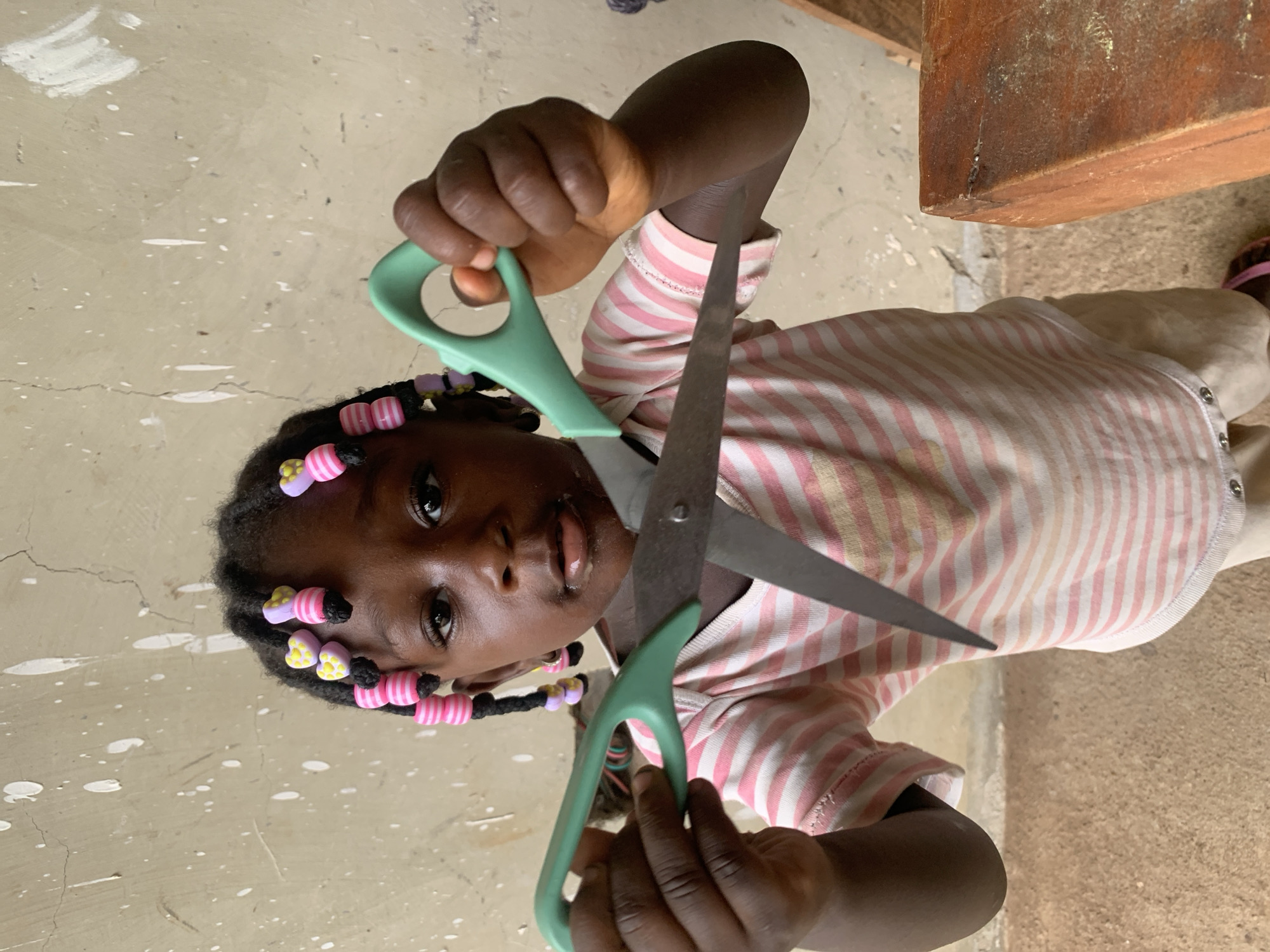 A child with dark skin and braided hair adorned with pink and purple beads is holding a pair of open scissors with green handles, directly facing the camera. The child is wearing a pink and white striped shirt. The scene appears to be indoors with a textured, light-colored floor. Part of a wooden structure is visible in the background. The location is Akwanga, Nigeria.