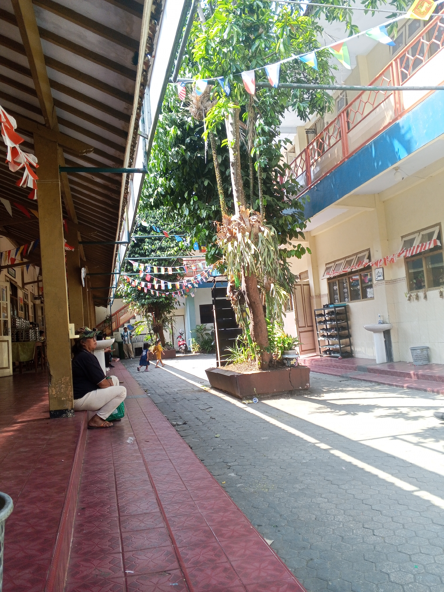 The image depicts a sunny outdoor courtyard with a covered walkway on the left. The walkway's roof is supported by wooden beams, and decorative flags are strung across the open space above. A person, appearing to be a woman wearing a black shirt, cream-colored pants, and sandals, is sitting on the red-tiled steps under the covered walkway, facing left. To her right, a green plastic container is visible. In the background, beyond a tree in a planter, a two-story building with blue trim and orange railings is present. Several windows with red and white striped decorations are visible on the building. A shoe rack holding multiple pairs of shoes, a sink, and a trash can are located outside the building. Two children, one in a blue dress and another in a yellow dress, are walking in the courtyard. The courtyard floor is paved with hexagonal gray stones. The scene appears to be in the city of Turen, Indonesia, indicated by a sign on the building that is partially visible.