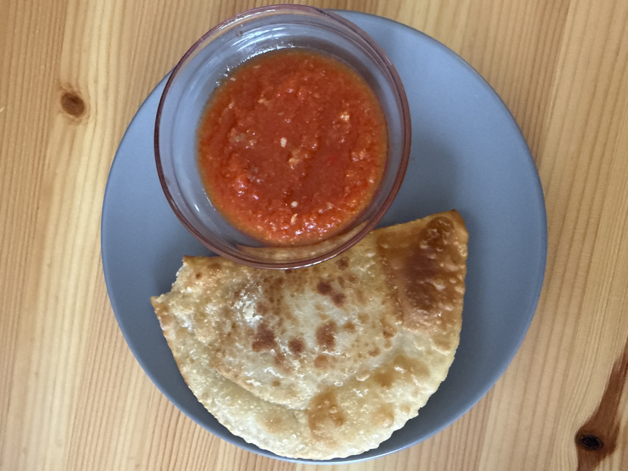 A top-down, close-up shot shows a gray plate on a light brown wooden table. On the plate, there is a small, clear glass bowl filled with a red sauce that appears to be made of pureed tomatoes and possibly spices or herbs. Next to the bowl, a golden-brown, fried pastry is placed. The pastry is folded and appears to be slightly crispy with some bubbly textures on its surface. The setting is casual, with the food arranged on a simple plate, suggesting a meal or snack. The lighting appears to be natural, possibly from a window, creating soft shadows and highlighting the textures of the food and the wood grain. No people are visible in the image. The context provided indicates the location is Zielona Góra, Poland.