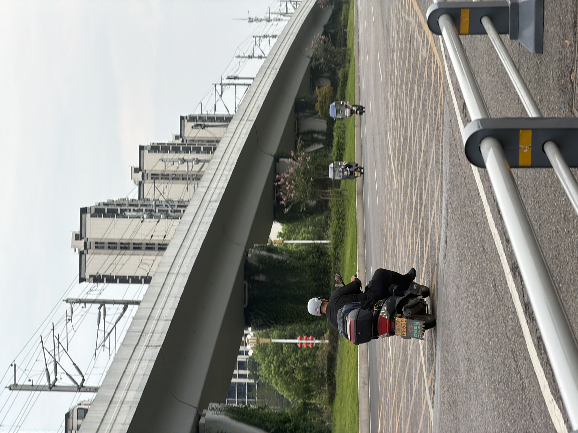 In Jiangjin, China, on an overcast day, a man wearing a black long-sleeved shirt, black pants, and a light gray helmet rides a black scooter. The scooter has a red taillight and a black storage box on the back, which contains a dark blue garment and a red item. The scooter's license plate is visible, reading "PF600". The road is asphalt with white and yellow lane markings. In the background, an elevated highway structure spans across the frame, supported by concrete pillars. Beyond the highway, a row of modern, multi-story buildings with dark windows is visible. Overhead, complex electrical lines associated with the elevated structure crisscross the sky. To the left of the road, green grass and trees line the embankment. Further down the road, two other scooters with canopies are visible, each carrying a rider. A silver metal guardrail with gray posts and yellow accents runs along the right side of the foreground.