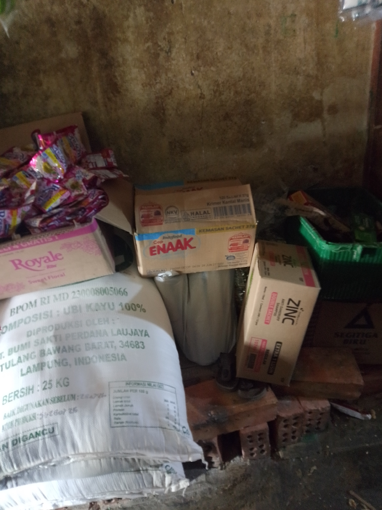 A collection of items, including food products and packaging, are arranged on a floor or surface, leaning against a textured wall. In the foreground, a large white sack is visible, with text printed on it in green ink, including "BERSIH : 25 KG" and "KEMASAN SACHET 37G". To the left of the sack, a pink box labeled "Royale" with "Sweet Floral" is partially obscured. Behind these items, a brown cardboard box with blue and white labeling, featuring the word "ENAAK" in large red letters, is present. To the right of the "ENAAK" box, another cardboard box, oriented vertically, displays the brand "ZINC" in white lettering against a pink background. A green plastic container is also visible in the background, filled with various items. The overall scene suggests a storage area or a retail environment, likely in Palembang, Indonesia, given the Indonesian text on the products. The environment appears somewhat cluttered, with bricks used to support some of the items. The wall behind the items is rough and textured, with signs of age or damage. No people are visible in the image.
