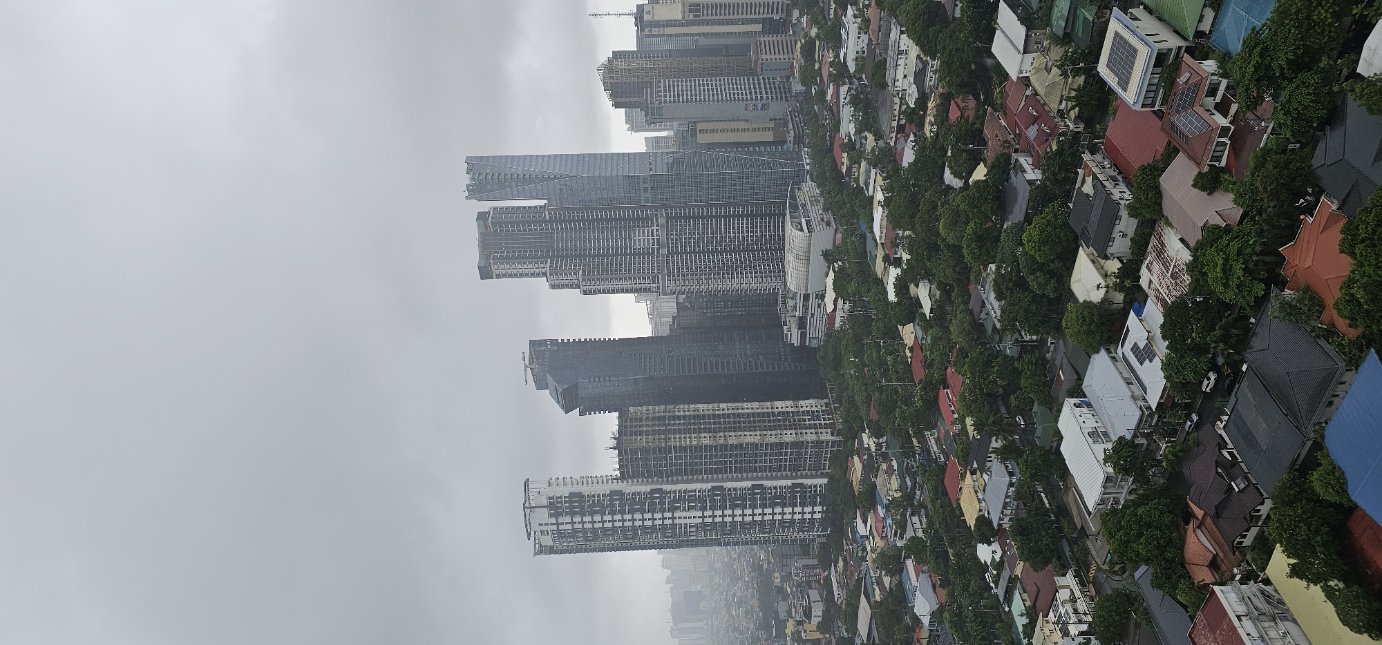 This is an aerial, vertical, full shot of a city scene under a cloudy, overcast sky. The scene is dominated by a dense urban landscape that transitions from modern skyscrapers in the background to a residential neighborhood in the foreground. In the background, a cluster of towering, contemporary skyscrapers, some with reflective glass facades, others with a more industrial, unfinished appearance, pierce the sky. Further in the distance, a hazy skyline suggests the vastness of the urban sprawl.
The midground is characterized by a more eclectic mix of buildings, including some mid-rise structures and what appears to be a large, rounded commercial building with a white, curved facade, possibly a mall or convention center. The foreground is occupied by a sprawling residential neighborhood, distinguished by a variety of houses with pitched roofs in various colors including red, brown, grey, and white. Lush green trees are interspersed throughout the neighborhood, creating a natural canopy that partially obscures the houses. Several vehicles are visible on the narrow, winding streets. The overall impression is one of a densely populated urban environment with distinct zones of commercial high-rise development and lower-density residential areas. The image appears to be taken in Makati, Philippines, given the characteristic architecture of the skyscrapers and the style of the residential buildings.