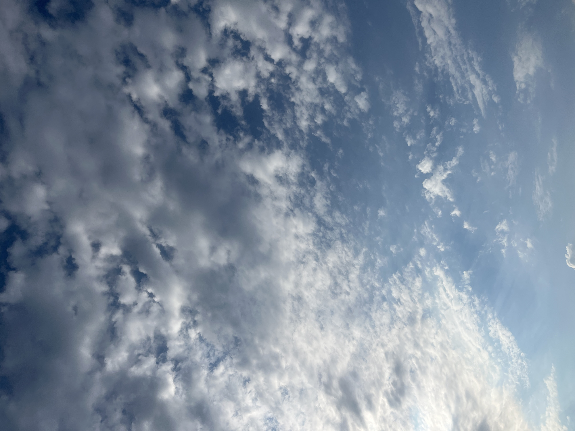 The image displays a sky filled with a mix of dark and light clouds against a blue background. The clouds appear to be in motion, creating a dynamic and textured effect. The lighting suggests it is daytime, with some areas of the sky being brighter than others. The scene is vast and open, with no discernible objects or structures on the ground, implying an outdoor setting, possibly a view from a high vantage point or an open landscape. There is no visible text in the image. The overall mood is natural and serene, with the interplay of light and shadow on the clouds creating a sense of depth and movement.