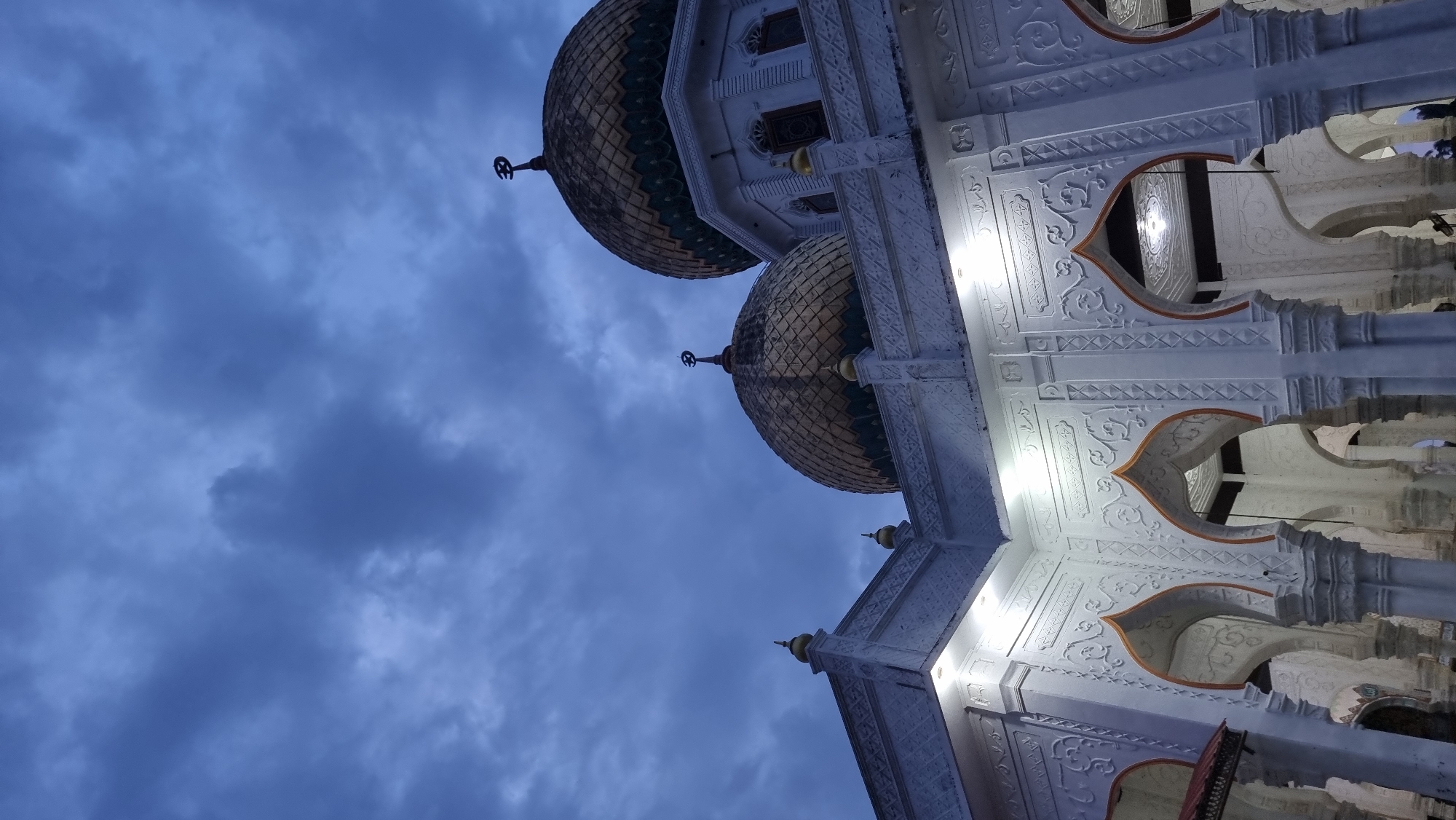The image shows the exterior of a mosque in Meunasahlhok, Indonesia, during twilight. The sky is overcast with dark blue and gray clouds. The mosque features white walls with intricate carvings and decorative arches. Two golden domes with green accents are visible at the top of the structure, each topped with a crescent moon. Lights illuminate the architectural details of the mosque. The lower portion of the image shows a series of arched colonnades.
