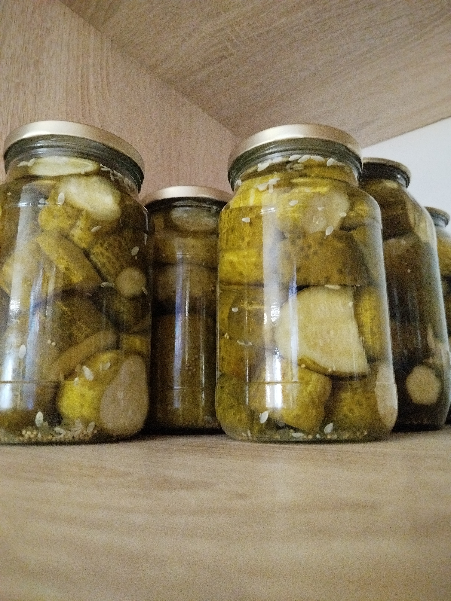 The image shows several glass jars filled with pickled cucumbers, likely homemade, arranged on a wooden shelf. The jars have metallic lids. The cucumbers are cut into pieces and appear to be submerged in brine, with some seeds and spices visible. The shelf is made of light-colored wood, and the background shows a similar wooden texture, suggesting a pantry or kitchen environment. There are no people visible in the image. The lighting is soft, creating reflections on the glass jars. The location in Zavodoukovsk, Russia, is not discernible from the image itself; this is external information.