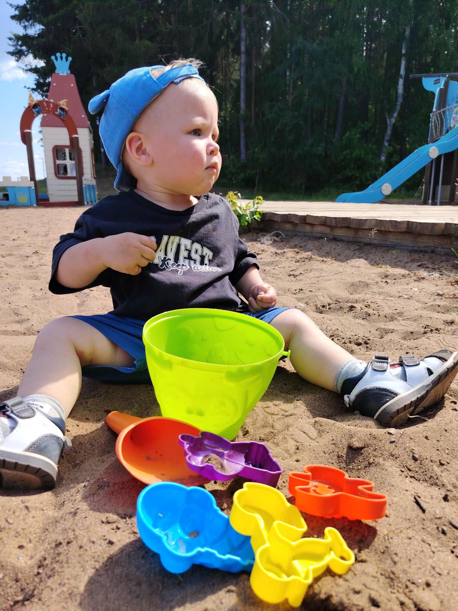 A young child, wearing a blue cap, a black t-shirt with white text that reads "WEST Keep active," and blue shorts, sits on a sandy playground. In front of the child are several colorful plastic toys: a bright green bucket, an orange shovel, and various shaped molds, including purple, blue, yellow, and orange ones. The child is looking to the right with a serious expression. In the background, a colorful playground structure resembling a castle and a blue slide are visible, set against a backdrop of tall, green trees. The ground is composed of sand, and the sky above is blue with some clouds. The scene appears to be an outdoor playground, possibly located in the city of Chaikovsky, Russia, given the context.