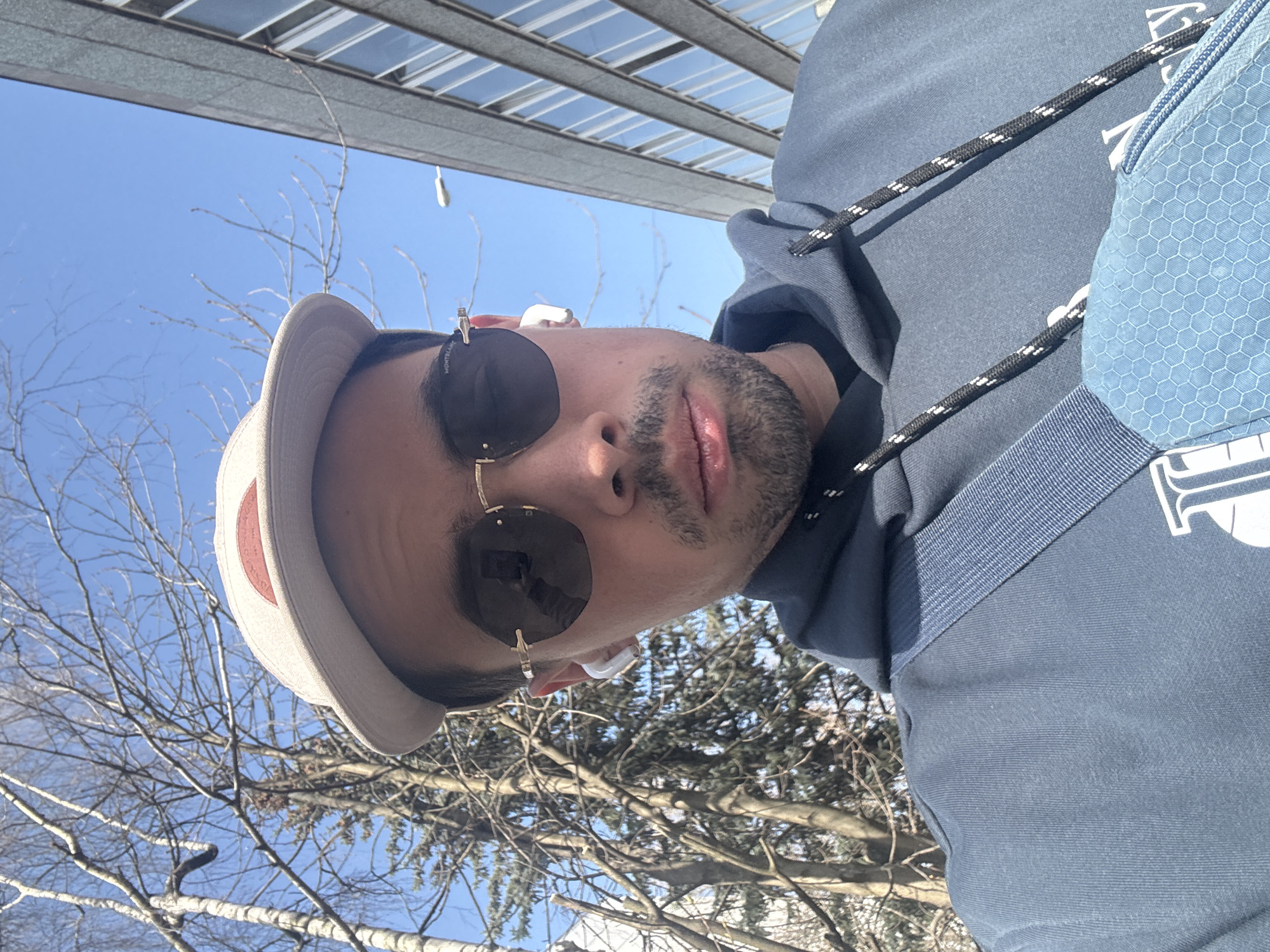 A low-angle, rotated headshot depicts a man wearing a beige hat, round sunglasses, and wireless earbuds, with a partial view of a blue hooded sweatshirt and a blue mesh bag. He is facing the camera with a neutral expression. The background features a blue sky, bare tree branches, and a building with a grid-like window pattern. The location is inferred to be Moscow, Russia, based on the architectural style of the building.
