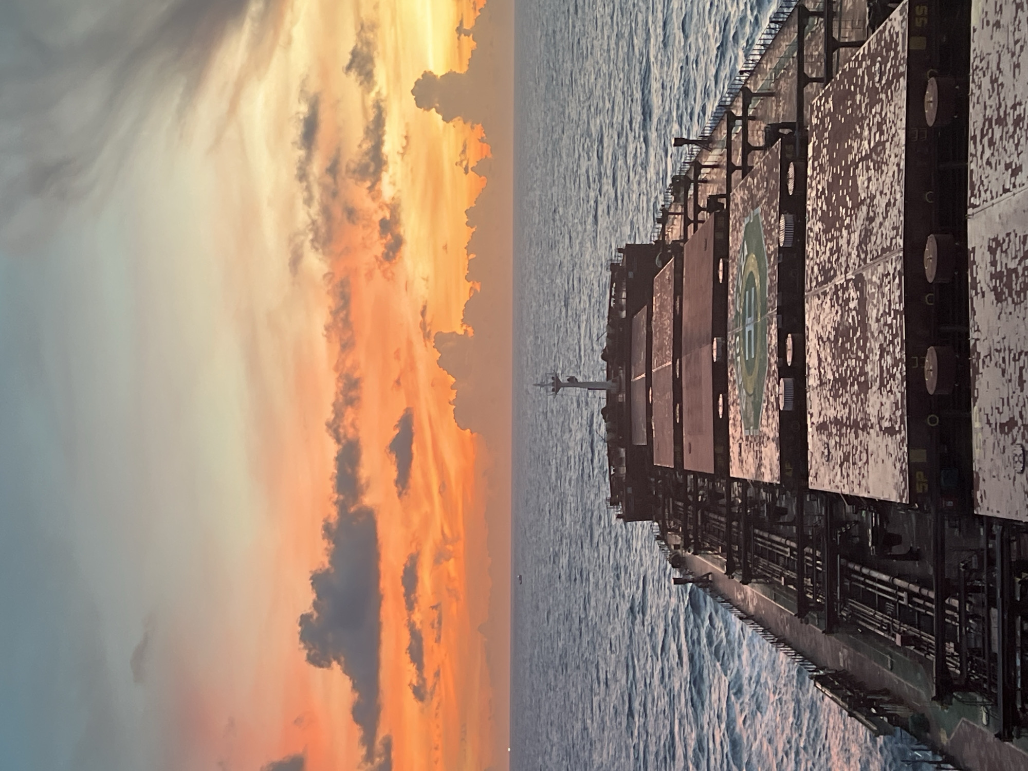 A perspective shot from the deck of a large cargo ship shows a dramatic sunset over the ocean. The sky transitions from a pale blue at the top to vibrant orange and pink hues near the horizon, with scattered gray clouds catching the light. The sea is a deep blue with gentle waves. The ship's deck is visible in the foreground, with numerous hatches and pipes. A helipad is visible on one of the hatches. The scene is tranquil and vast, emphasizing the isolation of the open sea. The image is taken at an unknown location in an unknown city.