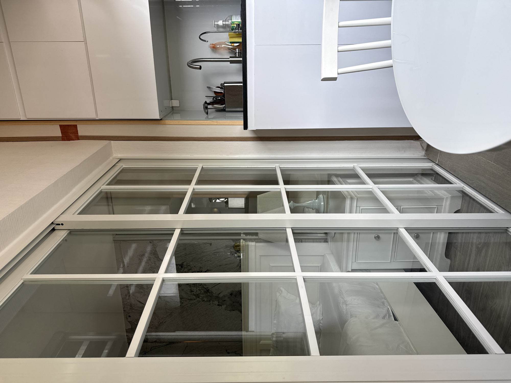 This image captures an indoor scene, split into two distinct areas separated by a large white-framed glass partition or window with multiple panes.
The upper section of the image (after correcting for the sideways orientation) reveals a modern kitchen or dining area. White, flat-panel cabinets line the wall. Beneath them, a stainless steel faucet with a pull-out spray is positioned over a sink, with dish soap, a sponge, and other cleaning items organized on a caddy. To the right, part of a round white dining table and a white chair with a ladder-style backrest are visible. A narrow strip of light wood trim and a hint of reddish wood trim separate the kitchen area from the glass partition.
The lower section of the image, viewed through the glass partition, shows another interior space, likely a bedroom or living area. Reflected light and the multiple windowpanes slightly obscure the view. Visible through the glass is a white cabinet with two drawers and circular silver pulls, topped by a decorative white lamp with a sculpted base. Further into this room, a large item, possibly a bed or sofa, is partially covered in white plastic sheeting, suggesting it might be new, recently delivered, or protected. The floor in this room appears to be a dark wood or wood-look tile. The walls are light-colored, possibly white, with some subtle paneling.
The overall aesthetic is bright, clean, and modern, with a dominant white color palette complemented by light wood accents and dark flooring in the distant room. No people are present, and no specific activity is taking place. There is no visible text in the image. The scene appears to be well-lit, likely by natural daylight.