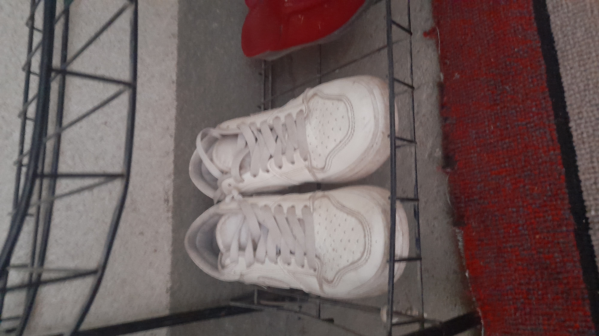 In a dimly lit indoor space, possibly a shoe rack or shelving unit, a pair of white low-top sneakers with light grey laces are prominently displayed. The sneakers appear to be well-worn, with visible creasing and scuff marks on the white leather exterior. They are positioned side-by-side on a metal shelf. To the right of the sneakers, partially visible, is a bright red object, possibly another shoe or accessory, also on the shelf. The background consists of a textured grey wall and a dark metal structure of the shelving unit. Below the shelf, a textured red rug is visible, with a darker border along the bottom edge. The image's quality is somewhat grainy, suggesting it was taken with a low-resolution camera or in poor lighting conditions. The overall scene appears to be a casual, everyday setting. The location is identified as Osh City, Kyrgyzstan, though the specific context within the city is not apparent from the image itself.