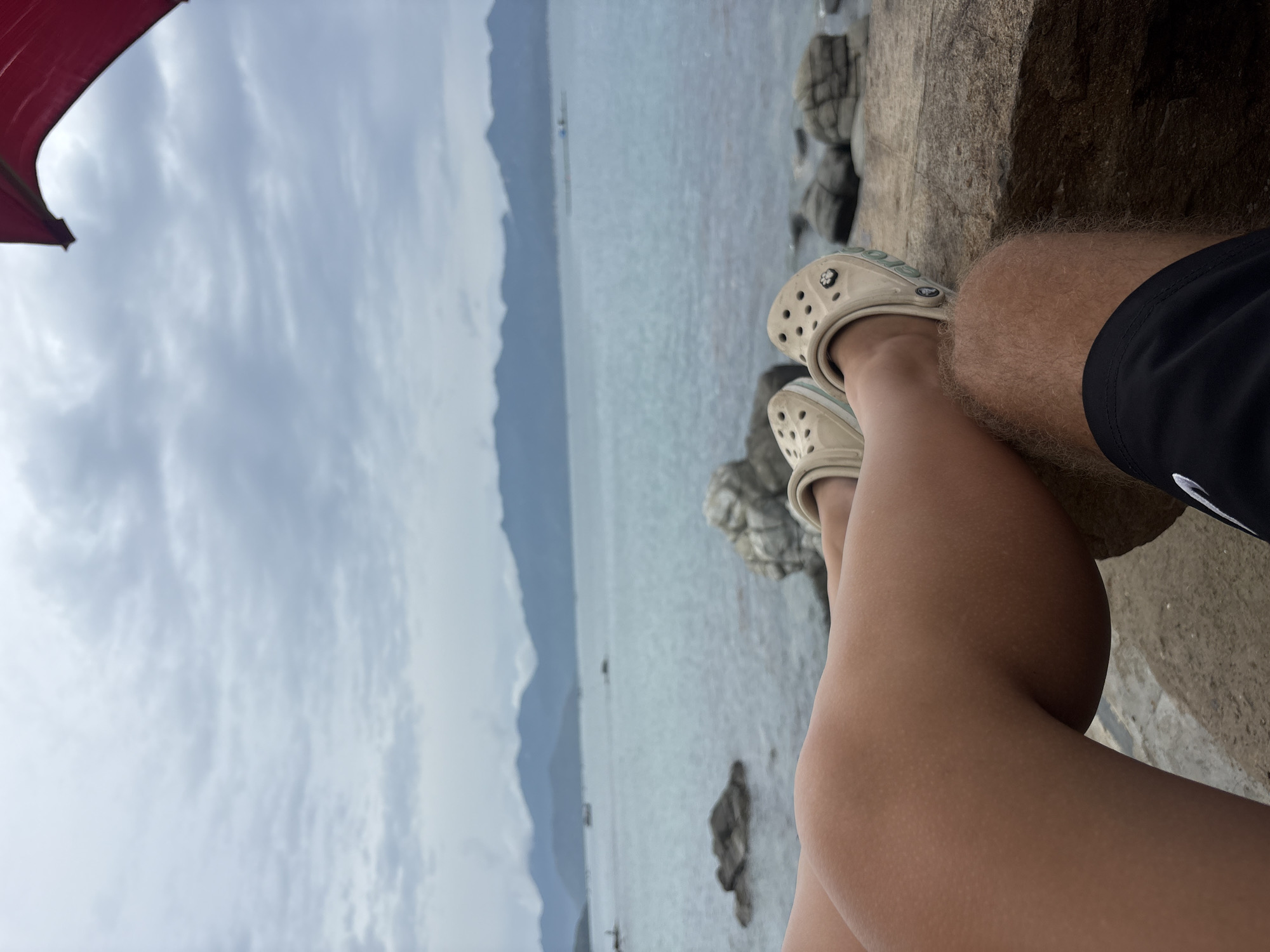 The image captures a first-person perspective looking down towards the viewer's legs and out over a body of water on an overcast day, set in what appears to be Vĩnh Lương, Vietnam. The shot is oriented vertically, with the horizon line running almost perpendicularly across the frame.
In the foreground, occupying the bottom right portion, are the legs of two individuals. One leg, with smooth skin, is seen wearing a light beige pair of Crocs clogs. A small, dark charm or logo, possibly a floral or star shape, is attached to the side of one Croc. Adjacent to this, the other leg is covered in fair-colored hair and is wearing dark (likely black) athletic shorts, with a small, partially visible white logo or symbol near the hem. Both sets of legs appear to be resting on a rough, dark brown rock surface, suggesting they are seated on a rocky shore or bank.
The midground features a vast expanse of calm, greyish-blue water, hinting at a sea or large lake. Several smaller, dark rocks are visible near the water's edge and partially submerged. In the distance across the water, a few indistinct shapes, possibly small boats or buoys, can be seen.
The background reveals a range of hazy, blue-grey mountains or large hills, appearing soft and layered under the cloudy sky. The sky itself is entirely overcast, filled with soft, diffuse grey and white clouds, indicating a lack of direct sunlight. In the top left corner, a vibrant red fabric object, likely a portion of an umbrella or canopy, provides a splash of color against the muted sky.
The overall scene conveys a sense of tranquility and relaxation, with the individuals enjoying a peaceful moment by the water on a cool, cloudy day.