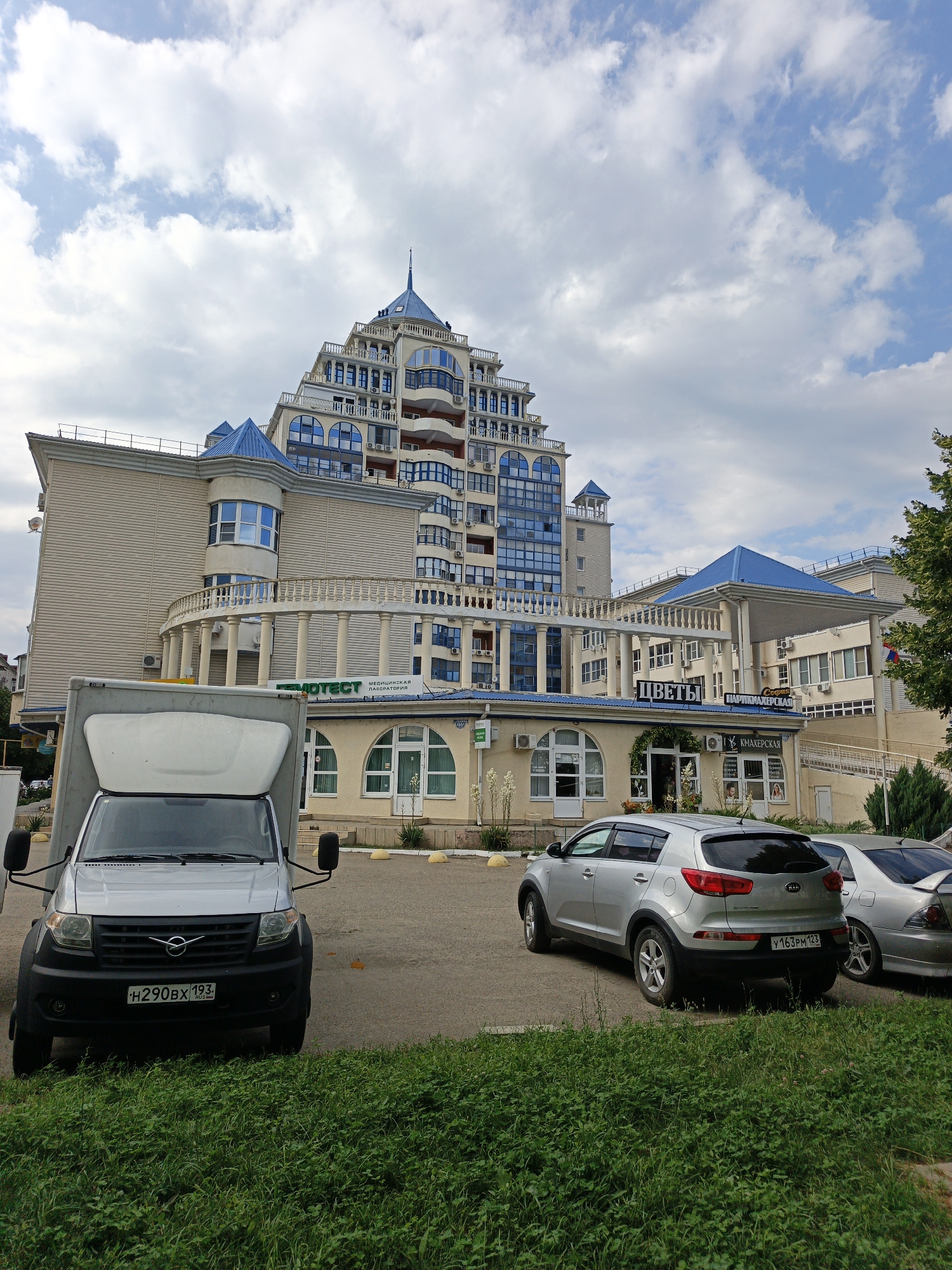 The image depicts a street scene in Goryachiy Klyuch, Russia, under a cloudy sky. The focal point is a modern, multi-story building with a distinctive pyramidal design and blue accents on its roof and windows. The building appears to house several businesses, with visible signage indicating "ФМOTECT МЕДИЦИНСКАЯ ЛАБОРАТОРИЯ" (FMOTECT MEDICAL LABORATORY) and "ЦВЕТЫ" (FLOWERS), along with "Студия ПАРИКМАХЕРСКАЯ" (Studio HAIRDRESSER) and "КНАЛЕРСКАЯ" (KNALERSKAYA), likely a salon.
In the foreground, a silver van with the license plate "H290BX 193" is parked. To the right of the van, a silver Kia Sportage SUV is parked, displaying the license plate "Y163PM 123". Another silver car is partially visible to the right of the Kia. The area in front of the building has a paved surface with yellow bollards and is bordered by a strip of green grass. The architecture of the building suggests a relatively modern construction, and the overall atmosphere appears calm, with no people visible in the immediate vicinity. The presence of shops suggests it is likely daytime, and the cloudy sky indicates the weather is overcast.