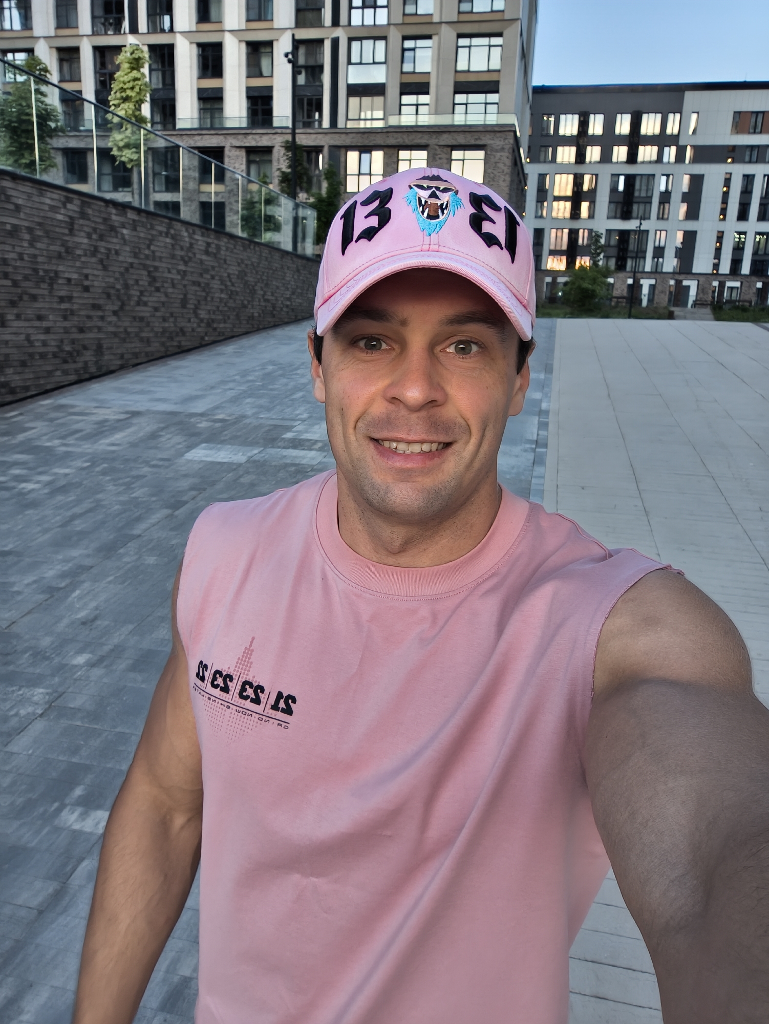 A man in a pink baseball cap and a pink sleeveless t-shirt is taking a selfie outdoors. The cap has the number "13" and a cartoonish blue lion's face on it. His t-shirt has a geometric pattern with black text. He is smiling at the camera. The background consists of modern apartment buildings, a brick wall, and a paved walkway. The lighting suggests it is either early morning or late afternoon. The location is the residential complex "Domodedovo Sky," located in Domodedovo, Moscow Oblast, Russia.