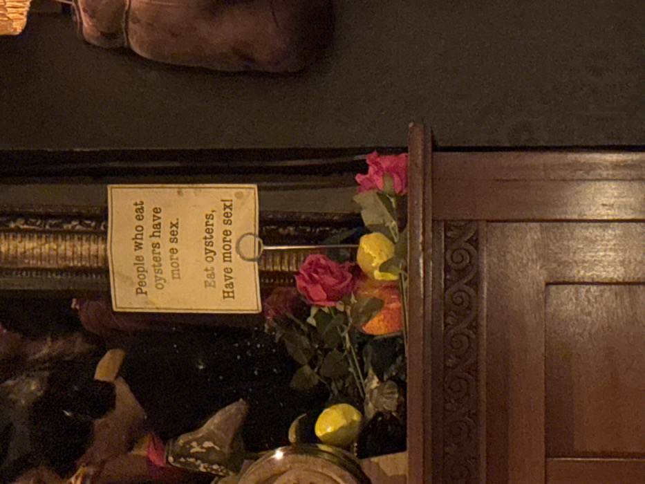 A humorous sign reads "People who eat oysters have more sex. Eat oysters, Have more sex!" It's displayed on a golden holder near a dark shelf, potentially in a restaurant or bar. Decorative elements including red roses and lemons accent the sign's placement. A dark wood frame with a carved design borders the scene. The photo's lighting is dim, creating an intimate, slightly mysterious atmosphere.