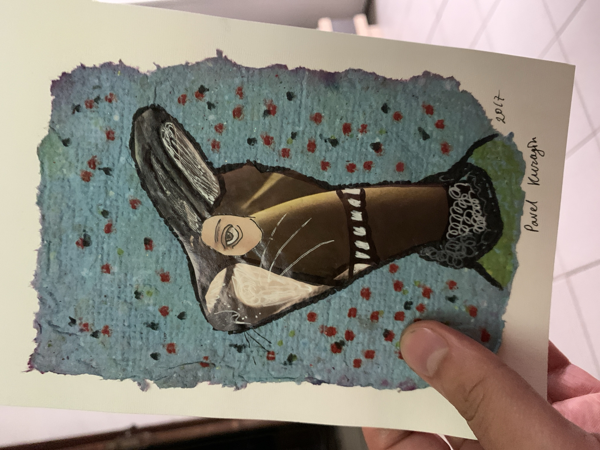 The image shows a postcard with a painting of a stylized creature with rabbit-like ears and a human-like eye. The background of the painting is a textured blue surface with scattered red and black dots. The creature has brown fur, a white snout, and a collar that resembles lace. The postcard is signed "Pavel Kuragin" and dated "2017" in black ink. A person's hand is holding the postcard, obscuring the lower left corner. The postcard is placed on a tiled floor. The image does not contain any information about the city of Sergeevo, Russia.