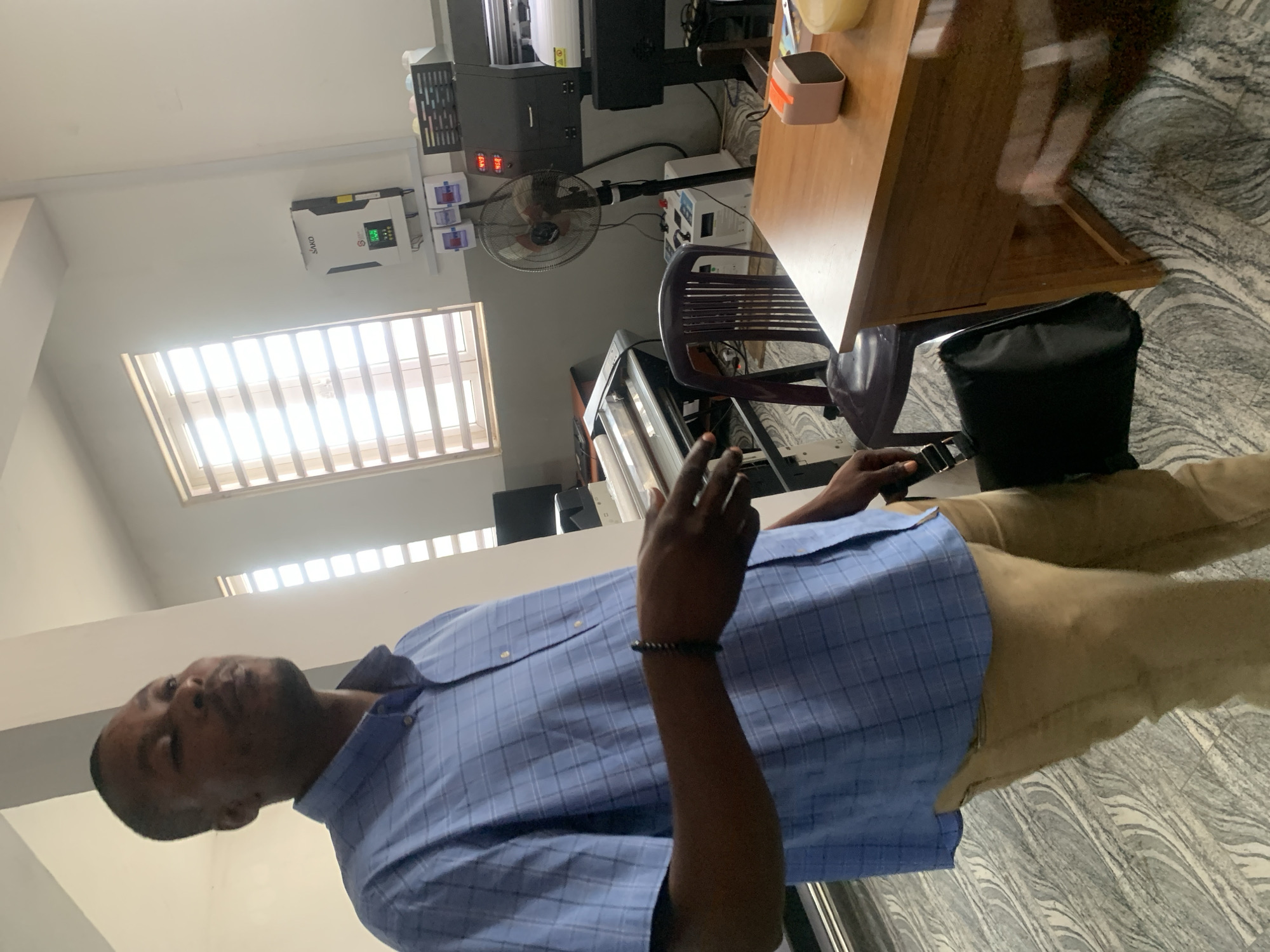 A medium shot shows a Black man in a blue checkered button-down shirt and tan pants holding a black bag in his right hand while standing indoors. He is facing left and looking up and away from the camera. His left hand is slightly raised, with his index finger extended. The man is in the foreground, and the background shows a room with windows, a desk, a fan, and some equipment. The room has gray and white patterned flooring.