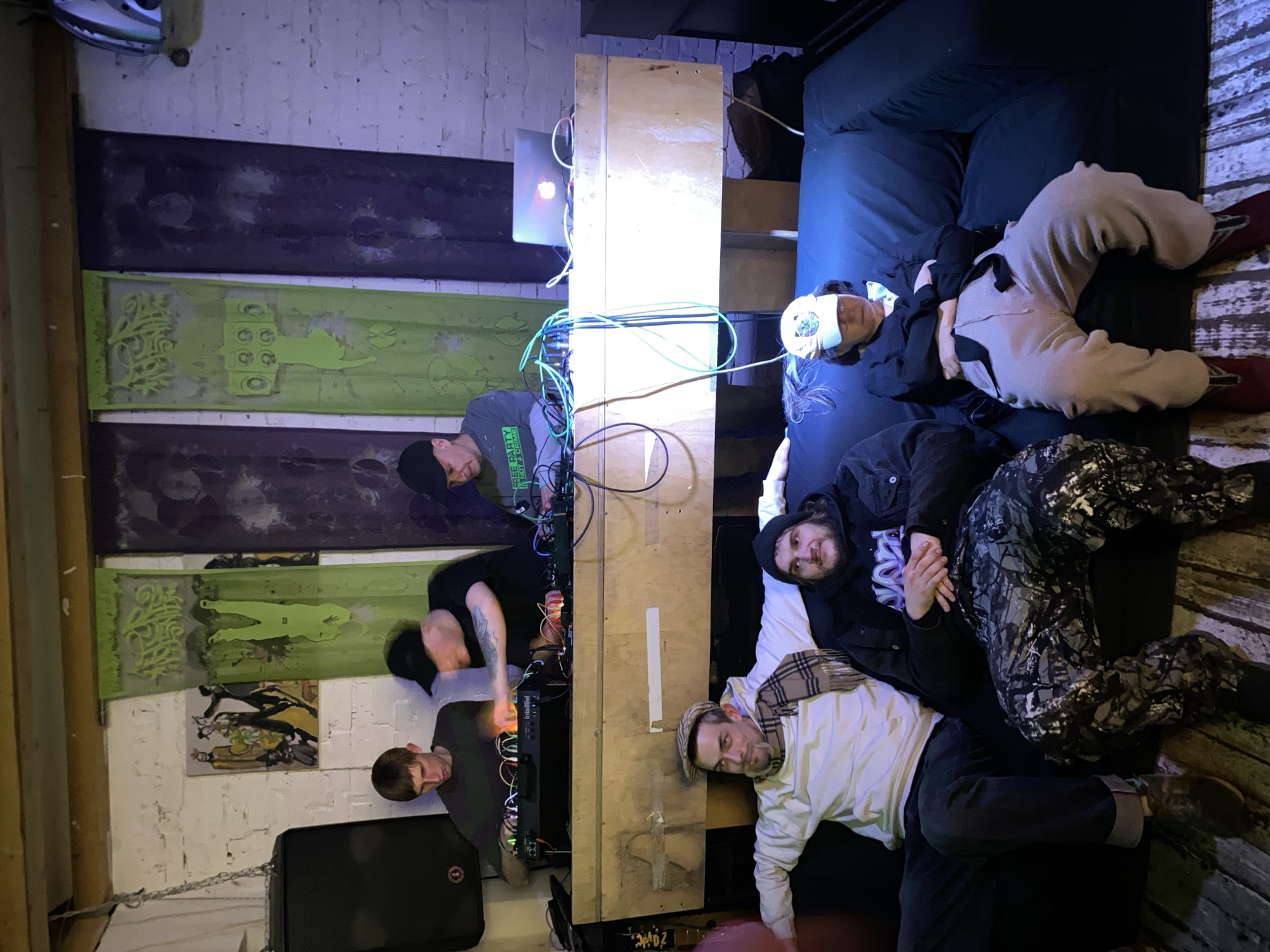 An indoor scene depicts multiple individuals engaged in a relaxed, possibly musical, activity. On the right, three people are reclining on dark cushions or mattresses. One person closest to the center has a white blindfold over their eyes, wearing a dark top and light-colored pants. Next to them, another person in a dark beanie and jacket with a plaid scarf lies on their side. Further right, a third person in a black jacket and camouflage pants also reclines.
In the center and left, three other individuals interact with a setup of electronic music equipment arranged on and around a tall wooden structure. Cables, including green, white, and blue ones, are prominent. A laptop with an illuminated Apple logo is visible near the top of the wooden console. Modular synthesizers, with "Intellijel" visible on one module, are being operated by the individuals. One person on the left wears a green shirt with "FREE PARTY" printed on it and a black beanie, bending over the equipment. Another person with tattoos on their arm is also focused on the gear, and a third person in a grey shirt observes the setup from a lower position.
The walls are painted white brick, partially covered by decorative panels: two dark purple/blue, and one bright lime green panel featuring intricate etched designs, including organic shapes and abstract figures. A small framed artwork hangs below the green panel. A black speaker is visible in the bottom-left corner, chained to a support beam. The lighting is dim overall, with a bright vertical light strip illuminating the central wooden console and casting a glow on the faces of those near it. The atmosphere suggests an intimate music production, listening, or jam session taking place in the evening or night.