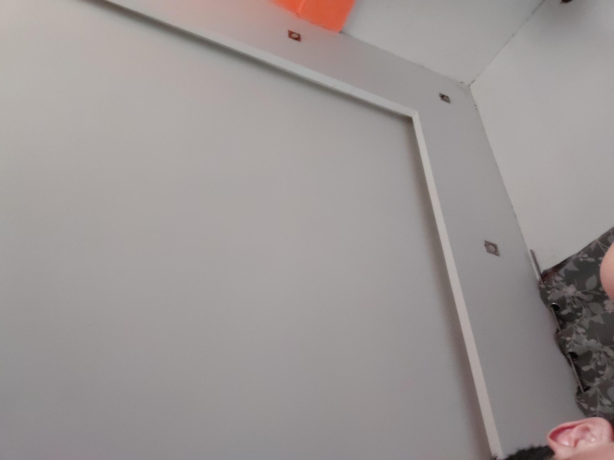 The image captures an indoor scene, likely a room within a home. The prominent features are light gray walls, with a large section on the left framed by a clean white trim. An orange object is partially visible in the top left corner, possibly a shelf or part of a ceiling fixture. Several small, dark, square-shaped elements are mounted on the walls and ceiling, which could be hooks, small speakers, or security camera mounts. The ceiling is white.
On the right side, a dark gray fabric with a subtle floral or leafy pattern is visible, suggesting a bedspread, curtain, or cushion. In the bottom right corner, a person's head, specifically their ear and a portion of their hair, is visible, along with a hint of a pink object below it, possibly a piece of clothing or an item on furniture. The photograph is taken from a low, tilted angle, looking upwards. There is no discernible text within the image. The time of day and specific weather conditions cannot be determined from this indoor view. No distinct location cues for Santa Ana, Venezuela, are present.