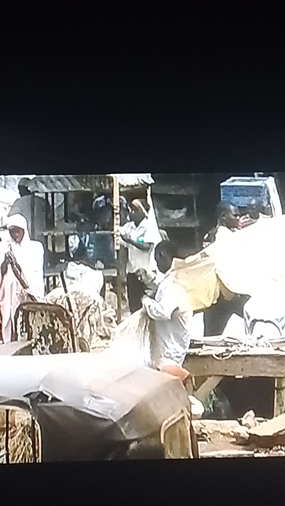 The image depicts a bustling market scene in Lokoja, Nigeria. Several individuals, predominantly men, are engaged in various activities. One man on the left, dressed in a white garment and headwrap, is standing with his hands clasped, seemingly in prayer or contemplation. In the center, a man in a light-colored shirt is interacting with someone behind a makeshift stall, possibly a vendor. Further to the right, another man in a white outfit is seen carrying a large, light-colored object, possibly goods for sale or transport. The environment appears to be an open-air market with wooden stalls and various structures in the background, suggesting a typical Nigerian urban setting. The ground is uneven, with visible debris and signs of daily activity. The overall impression is one of lively commerce and community interaction.