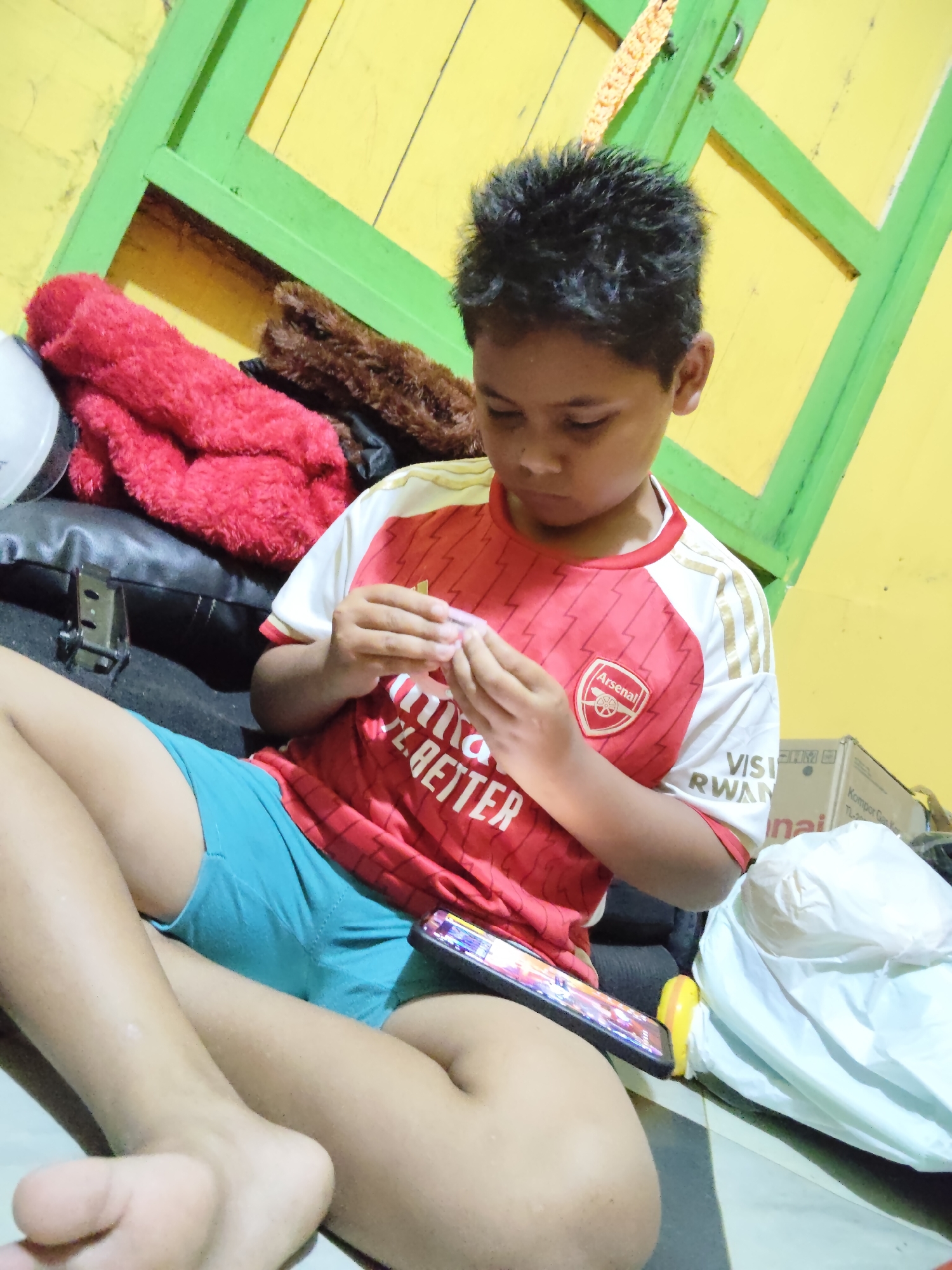 A young boy, appearing to be of Southeast Asian descent, is seated on the floor in an indoor setting. He is wearing a red and white Arsenal football jersey and blue shorts. His hair is dark and spiky. The boy is looking down at his hands, which are holding a small, pink object, and appears to be concentrating on it. A smartphone, displaying a colorful game interface, is placed on his lap.
The environment appears to be a room with yellow walls and a green-painted wooden frame or door. To the left of the boy, there are several items on what looks like a black leather surface: a white helmet, a red fuzzy blanket or towel, and a brown fuzzy material. In the background, there are cardboard boxes and white plastic bags, suggesting a storage area or a room with discarded items. The floor is tiled. Based on the overall appearance and common cultural motifs, this scene is consistent with a residential interior in Tanah Grogot, Indonesia.