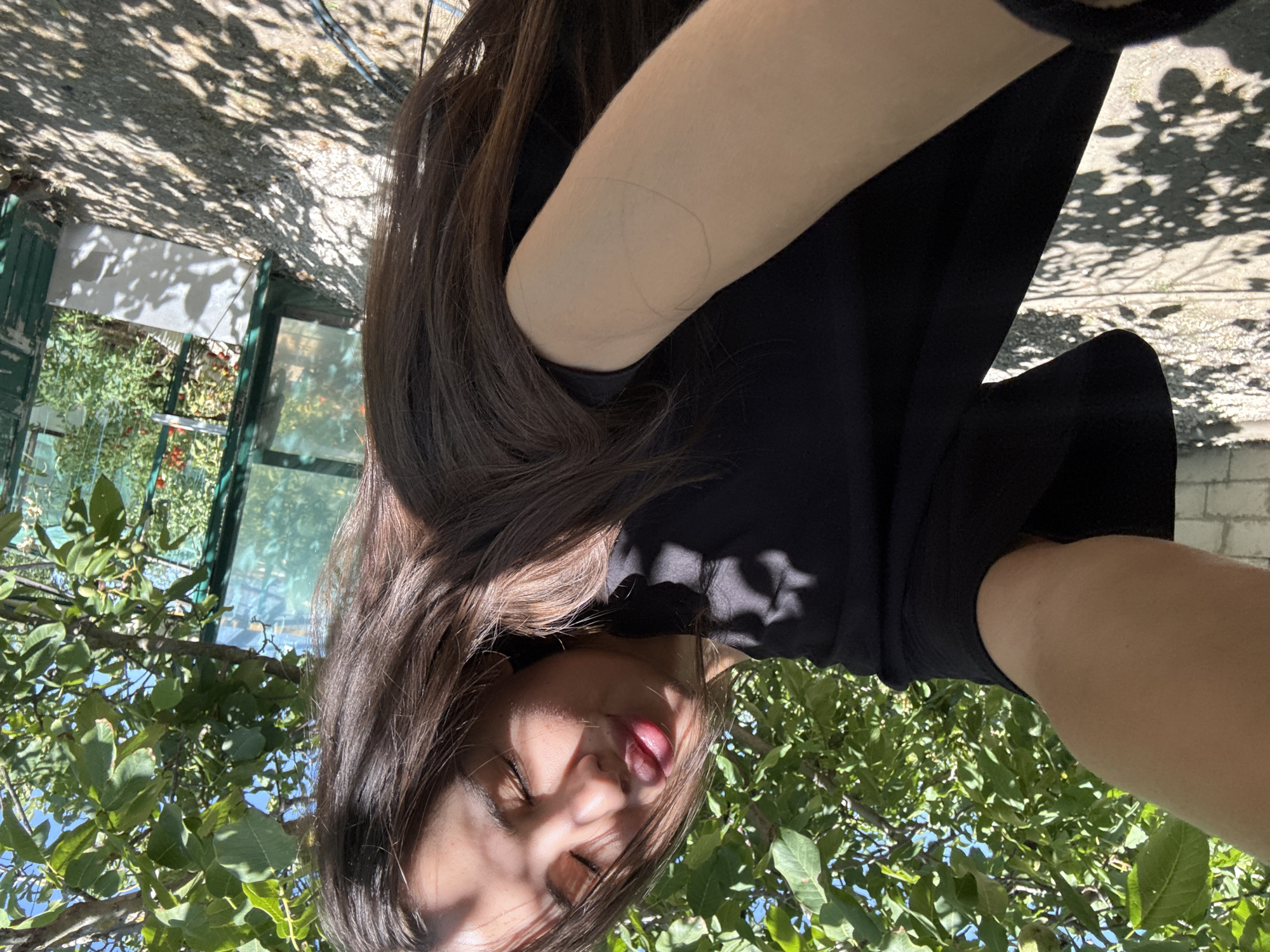 The media file is a selfie taken by a person outdoors, with their face and arm visible. They are wearing a black t-shirt. The scene is set in a garden or yard, with lush green foliage from trees and bushes surrounding the subject. A greenhouse structure with green framing and glass panels is visible in the background, containing rows of plants, possibly tomato plants with red fruit. Sunlight filters through the leaves of trees, casting dappled shadows on the ground and the subject. The lighting suggests it is daytime, likely a sunny day, given the bright shadows. The subject's expression appears playful, with their eyes closed and tongue sticking out slightly. The image seems to have been taken from a low angle, looking up at the subject and the surrounding greenery. There is no visible text in the media file.