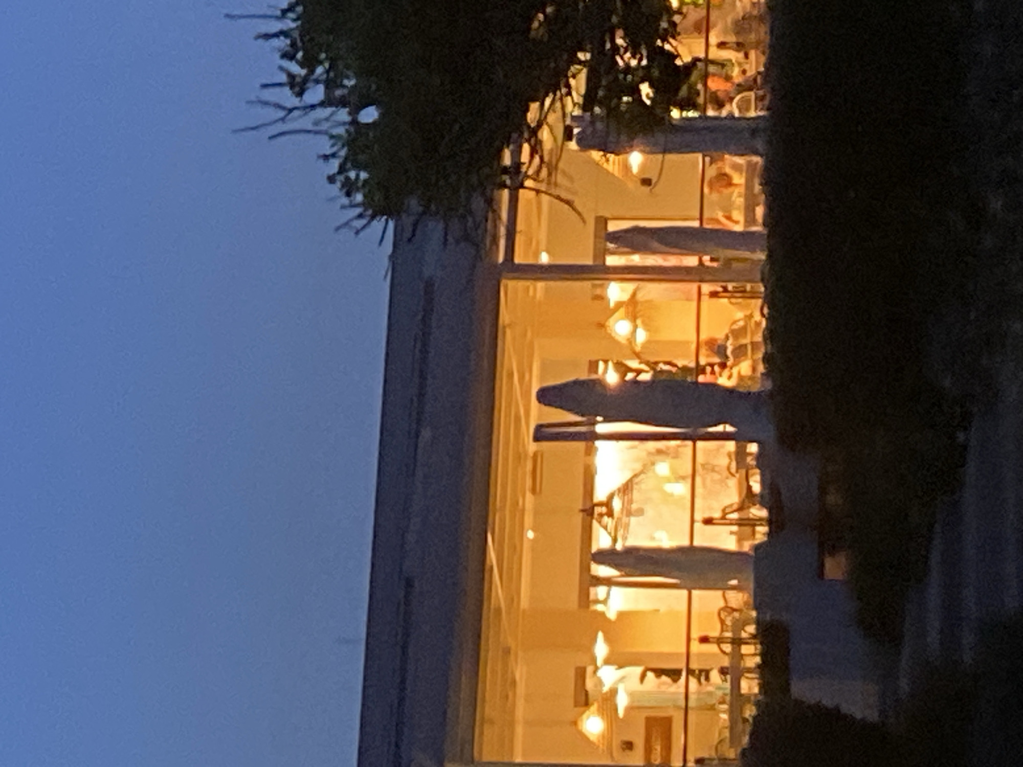 The image captures a dimly lit scene at dusk, likely in Estepona, Spain, showing the exterior of a building with large windows, appearing to be a restaurant or cafe. Inside, warm interior lighting illuminates tables with patrons seated and engaging in conversation. Several tall, light-colored umbrella-like structures stand in the foreground, partially obscuring the view of the building's interior. A dark, dense tree is visible on the right side, its branches silhouetted against the twilight sky. The overall atmosphere is serene, with the building and its occupants bathed in a soft, inviting glow.