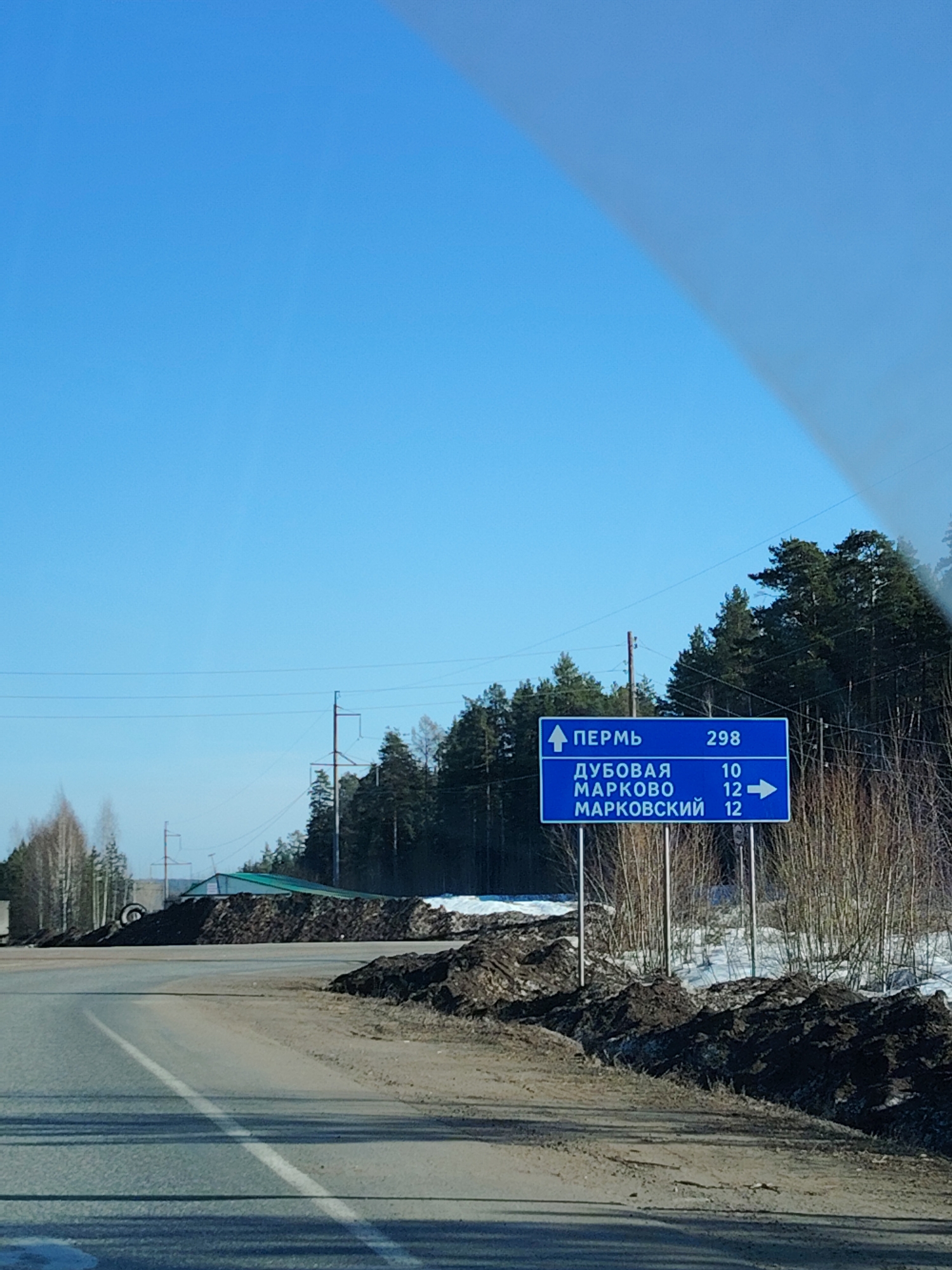 The image shows a road scene in Chaikovsky, Russia, with a blue road sign in the foreground indicating distances to Perm (298 km), Dubovaya (10 km), Markovo (12 km), and Markovsky (12 km). The sign has a directional arrow pointing forward for Perm and an arrow pointing right for Markovo and Markovsky. The road is asphalt with a white lane marking and has dirt on either side. A large pile of dirt and debris is visible on the left side of the road behind a building. In the background, trees are seen under a clear blue sky. Power lines are also visible. It appears to be a rural area or the outskirts of a town. Patches of snow are present.