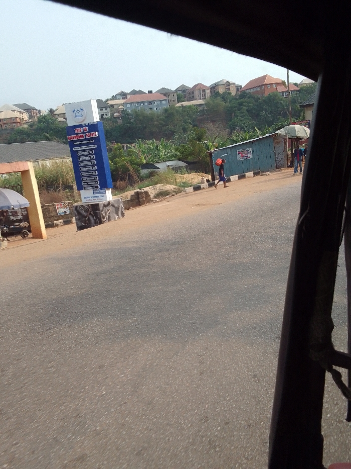 The scene depicts a daylight street view in Nkwelle Ezunaka, Nigeria, captured from within a vehicle. The asphalt road surface occupies the foreground and mid-ground. To the left, a light-colored archway stands next to a parked motorcycle under an awning. Centered mid-left, a prominent blue and white vertical sign displays "THIS IS RIVERBANK ESTATE" along with a list of addresses. To the sign's right, a dirt shoulder with black and white striped curbing borders the asphalt. On this shoulder, a small, blue corrugated metal shack is visible, along with another structure partially obscured by an umbrella. One individual, wearing dark shorts and a shirt, walks rightward on the dirt shoulder, carrying an orange container on their left shoulder. Two additional figures are indistinctly visible near the blue shack. The background features a densely vegetated hillside dotted with numerous multi-story residential buildings exhibiting various roof colors (e.g., red, brown, grey). Dark structural components of the viewing vehicle frame the top and right edges of the image.