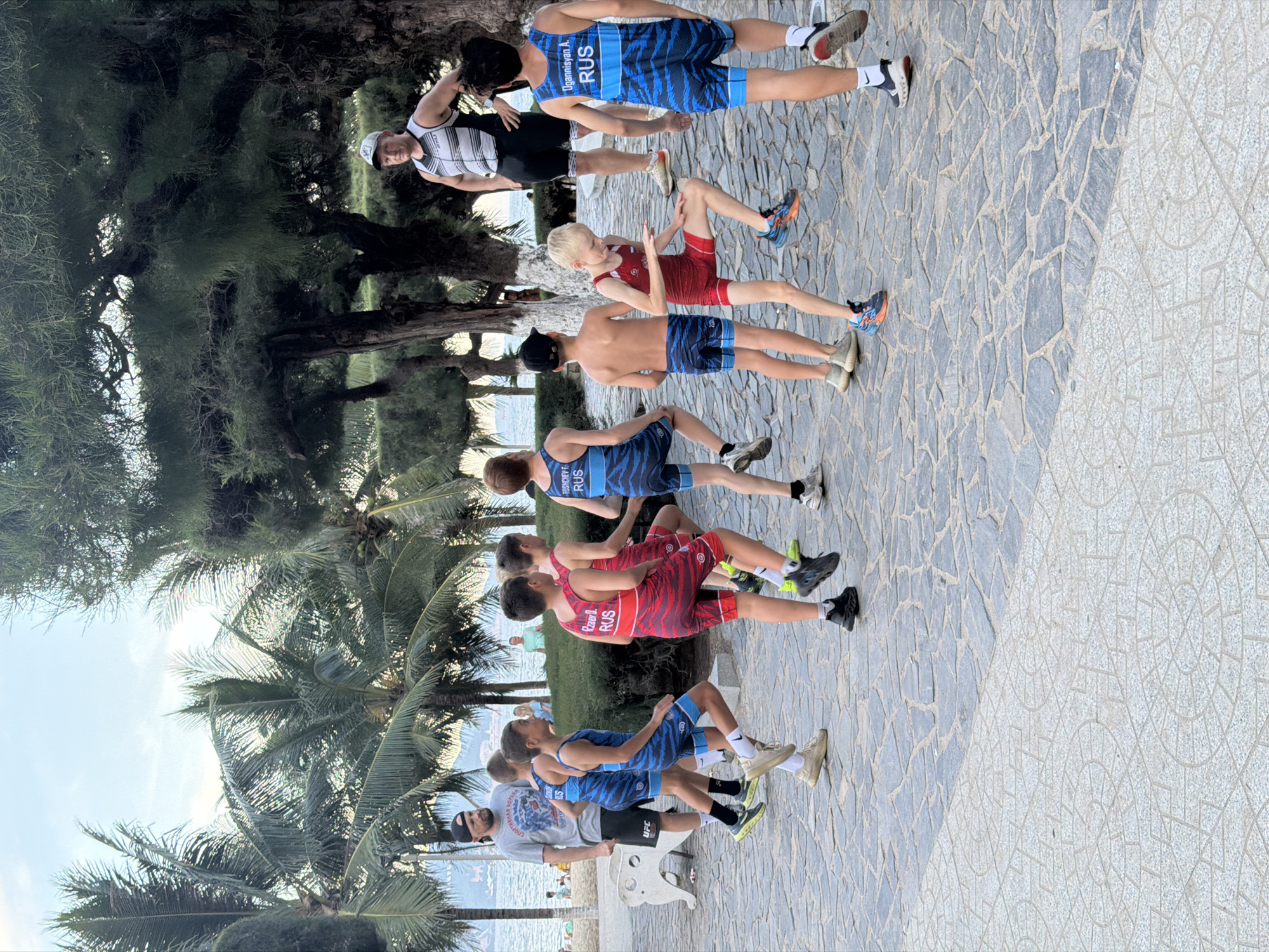 The image shows a group of people, likely wrestlers, posing for a photo in Nha Trang Ward, Vietnam. The subjects are arranged in a line on a paved walkway, with palm trees and lush foliage in the background, suggesting a tropical setting. The time of day appears to be daytime, with bright lighting consistent with clear skies and warm weather.
The group consists of at least ten individuals, primarily young men and boys, dressed in athletic attire. Several are wearing wrestling singlets with the "RUS" insignia, indicating they represent Russia. The singlets feature blue and black tiger stripe patterns. One person wears a black and white striped tank top and shorts, and another a red wrestling singlet. Some individuals are wearing wrestling shoes, while others have athletic sneakers.
The individuals are in various poses, some standing, others seated or in a semi-crouched position. The overall mood seems relaxed and celebratory, as if they are documenting a team event or competition. In the background, the ocean and a distant coastline are visible, further reinforcing the beachside or coastal location. The paved walkway is made of grey stone tiles of varying sizes, with a section of lighter, patterned tiles to the right.
Visible text includes "UGANNISYAN A. RUS" and "RUS" on some of the singlets. One person in the foreground is wearing a grey t-shirt with a "UFC" logo. The scene suggests a team activity or team photo session in a pleasant, outdoor environment.