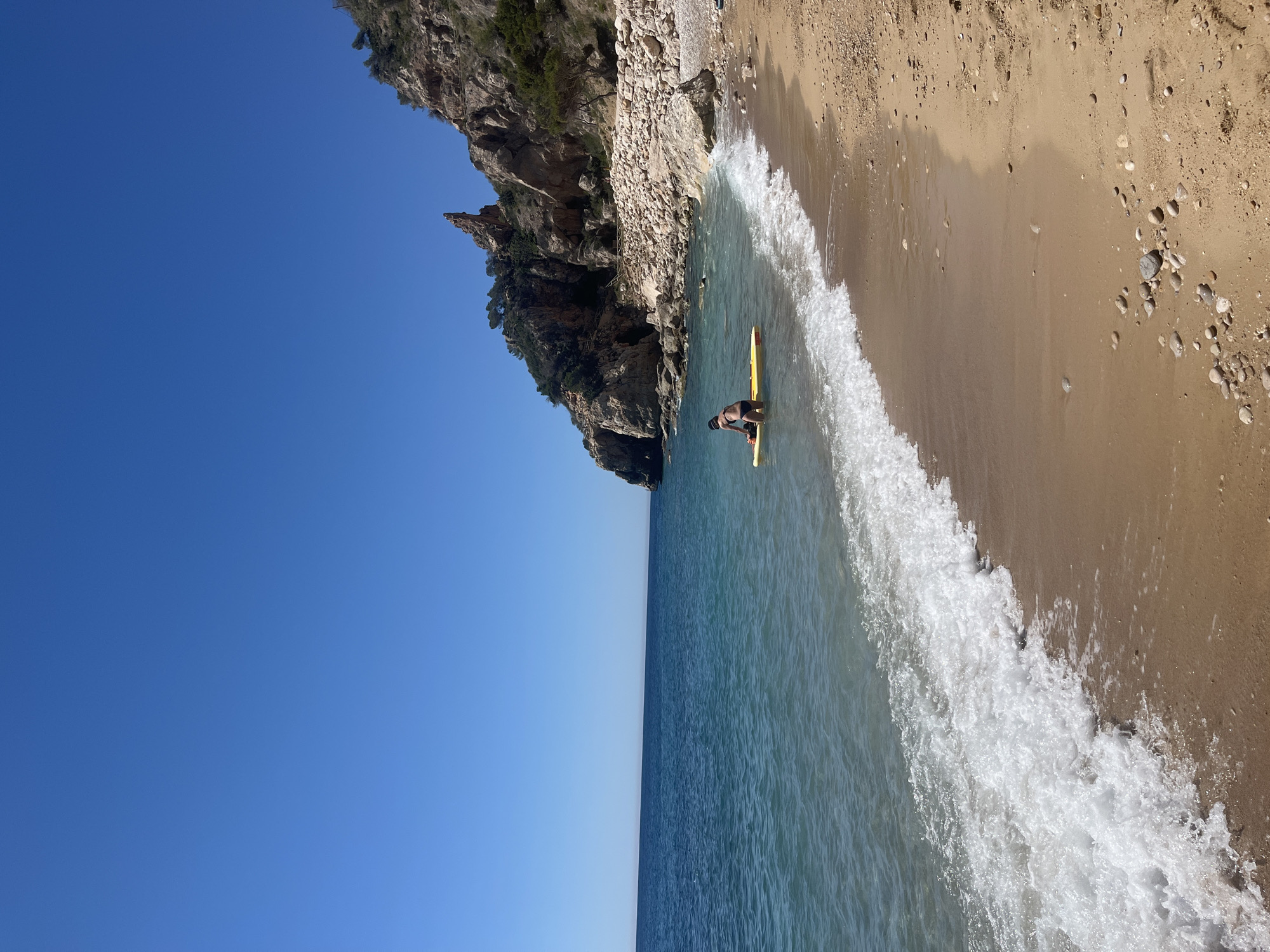 The image shows a beach scene at Cala del Moraig in El Poble Nou de Benitatxell/Benitachell, Spain. A rocky cliff rises on the left side of the beach. A person with a black cap and dark swimming trunks is standing in the water with a yellow surfboard. The water meets the sandy beach with foamy waves. The sky is clear and blue. The image is taken at an angle.