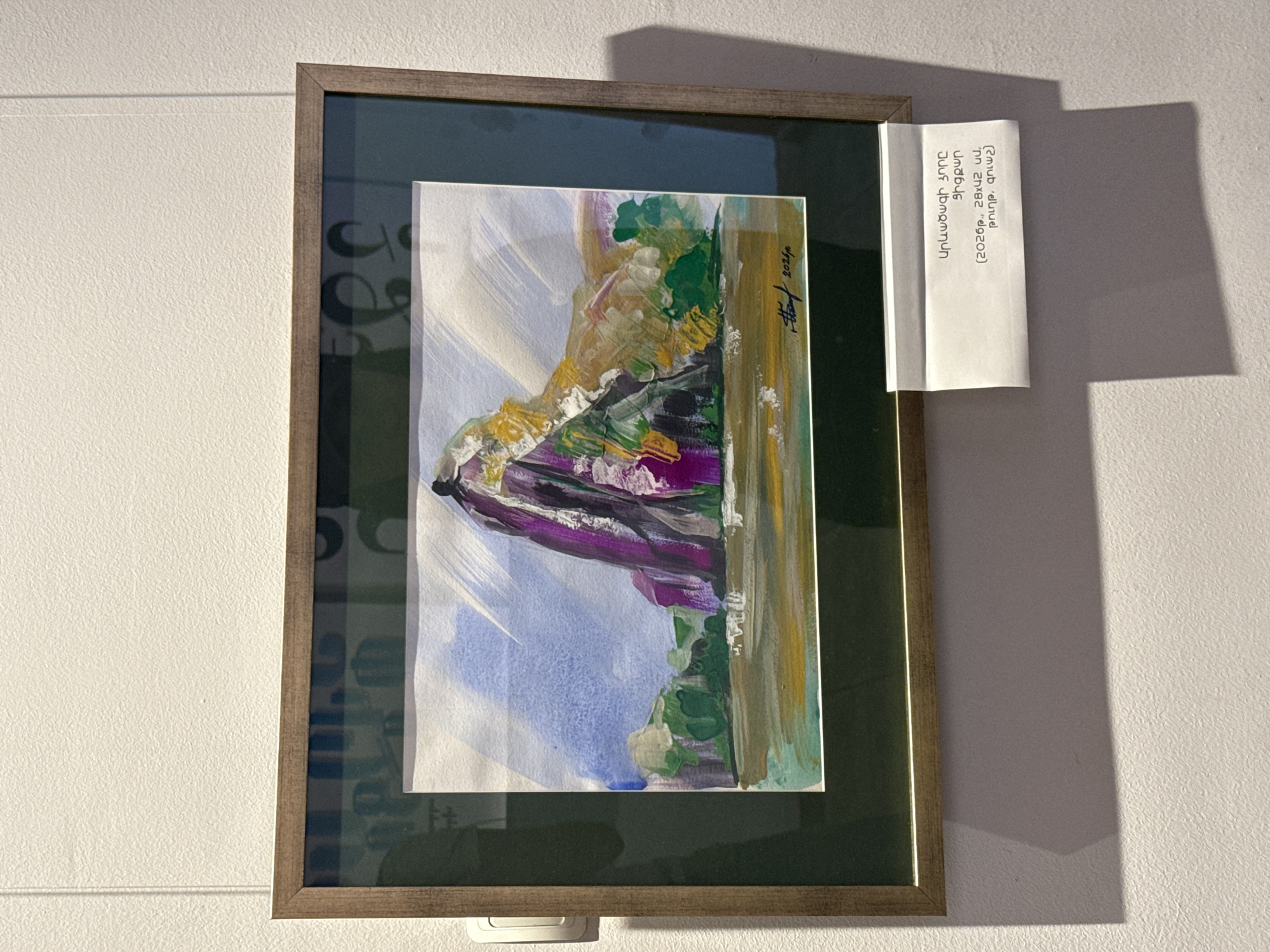 A framed painting hangs on a light-colored wall. The painting depicts an abstract landscape with purple, green, yellow, and blue colors. A white card with text in Armenian is attached to the right side of the frame. The text on the card includes "Արևածագի ձորը Ցիցքար (2026р., 28x42 ud, թուղթ, գուաշ)". The painting is signed with what appears to be "Hthon" and the year "2024". The frame is brown with a dark green inner border. A shadow is cast on the wall to the right of the frame. The location is Yerevan, Armenia.