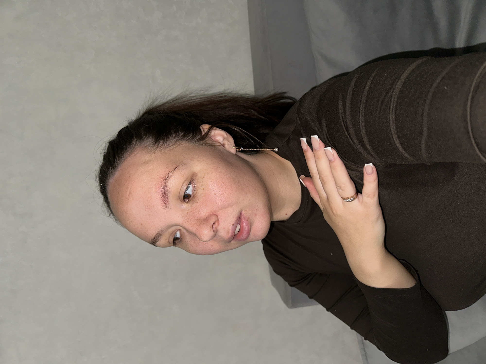A woman with brown hair is positioned in the lower left quadrant. She is wearing a brown long-sleeved shirt and a gold earring. Her right hand is raised near her face, showing nails with white tips and a gold ring on her ring finger. The background consists of a gray wall and a gray surface, possibly a sofa. The picture could be from Chaikovsky, Russia, as requested, but there are no visual indicators within the image itself to confirm this location.