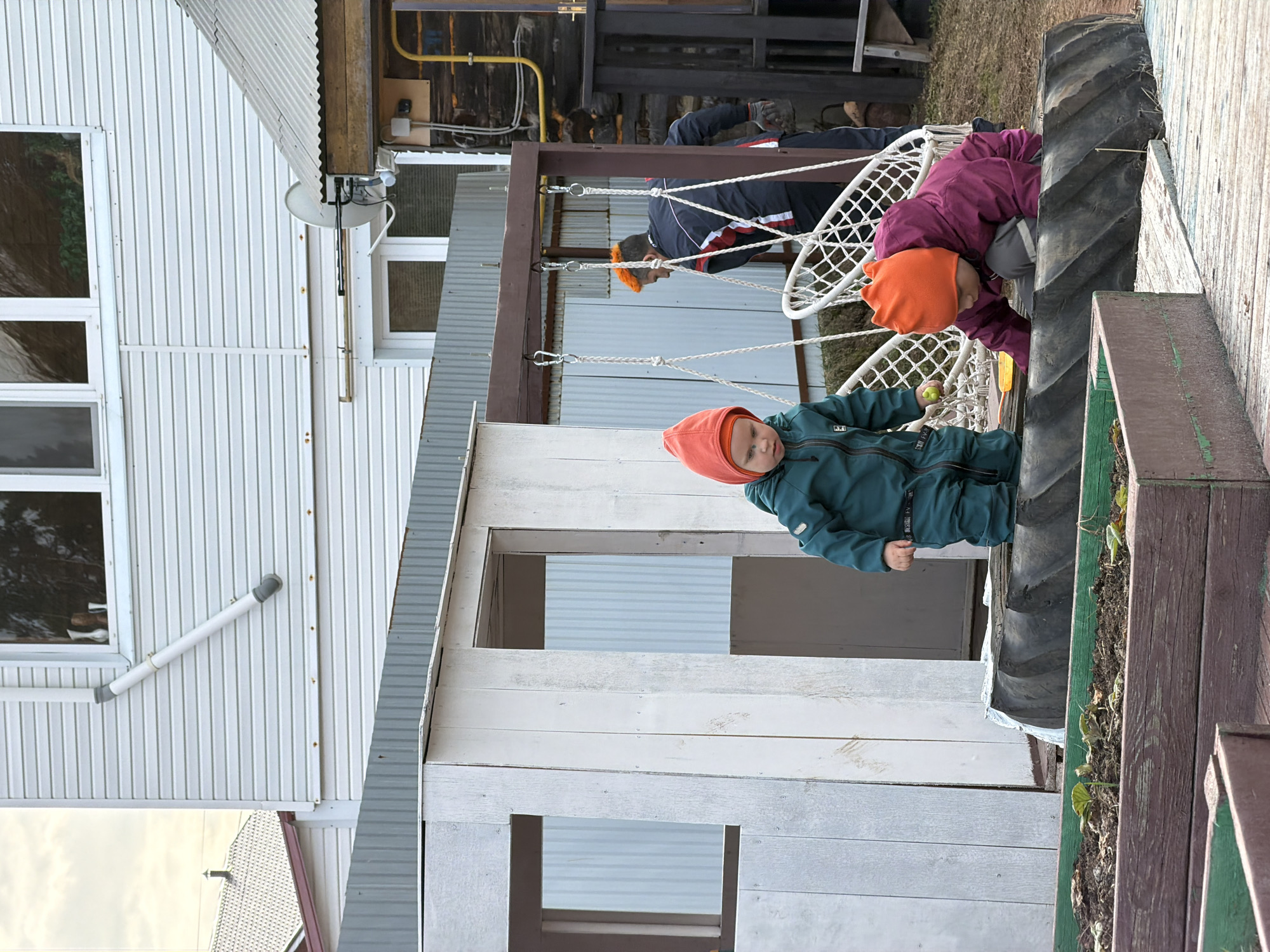The image depicts a residential area in what is likely Chaikovsky, Russia. There's a white siding building with multiple windows and a gray corrugated roof visible on the left. Attached to this is a small white wooden structure with what appear to be open window frames and a door.
In the foreground, a young child wearing a green jumpsuit and an orange hat is standing. Behind the child, there's a large tire used as part of the play structure, next to a wooden planter box. Further back, two people are relaxing in hanging hammock chairs suspended from a brown wooden frame. One person is wearing a dark blue jacket with white and red accents and an orange hat, while the other has on a purple jacket and an orange hat. There is also some type of yellow tubing on the wooden frame. The ground is covered in grass and some other outdoor items.