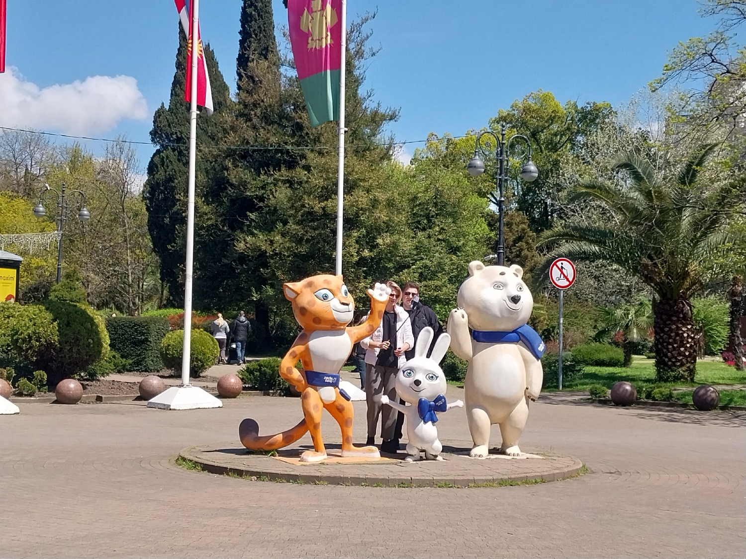 This vibrant outdoor photograph captures a sunny scene in Sochi, Russia, featuring the three official mascots of the 2014 Winter Olympics: the Leopard (Bardy), the Hare (Zaika), and the Polar Bear (Bely Mishka). The mascots stand prominently on a paved circular platform in the foreground. The leopard, an orange figure with dark spots, is on the left, wearing a blue sash. In the center is the white hare with a blue bow, and on the right stands the large white polar bear with a blue scarf.
Behind the mascots, two men and a woman are visible, appearing to pose for a photo with the figures. One of the men holds what looks like an ice cream cone. The setting is a lush park or promenade area, characterized by abundant greenery, including tall coniferous trees, various deciduous trees, and a distinct palm tree on the right, indicative of Sochi's subtropical climate. Two flagpoles stand tall behind the mascots, flying a white and red flag and a green and gold flag, respectively.
Notable details include a classic black street lamp, decorative dark spherical bollards lining the paved path, and a red circular prohibitory sign on the right depicting a crossed-out figure on a scooter or skateboard. Partially visible text on the mascots' sashes reads "Sochi 2014," and a yellow sign on the far left corner shows "maxim." The bright blue sky with a few white clouds indicates a clear and pleasant day. Several other people are seen walking in the background, adding to the lively park atmosphere.