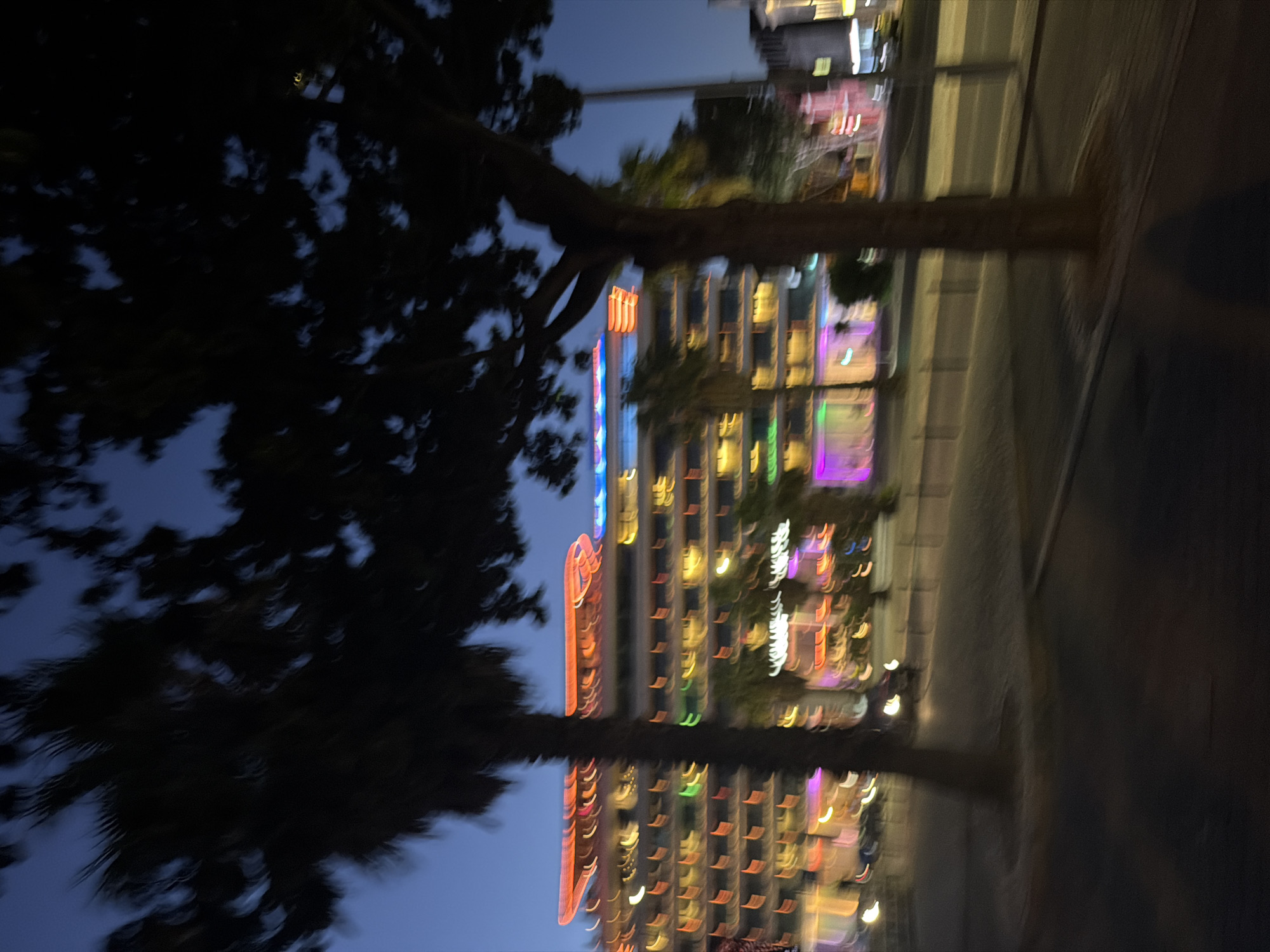 The image shows a blurred nighttime scene of a building with illuminated windows and colorful lights. A dark silhouette of a tree is in the foreground, partially obscuring the view of the building. The lights on the building are streaks of various colors, including orange, red, yellow, green, and purple, suggesting a vibrant urban environment at night. The sky is a dark blue, indicating dusk or nighttime. The reflection of the building and lights can be seen in what appears to be a body of water or a wet surface below, adding a symmetrical element to the composition. The overall impression is one of a lively city or resort area after dark. The motion blur suggests the photo was taken while moving, perhaps from a vehicle or on foot. There is no visible text in the image.
