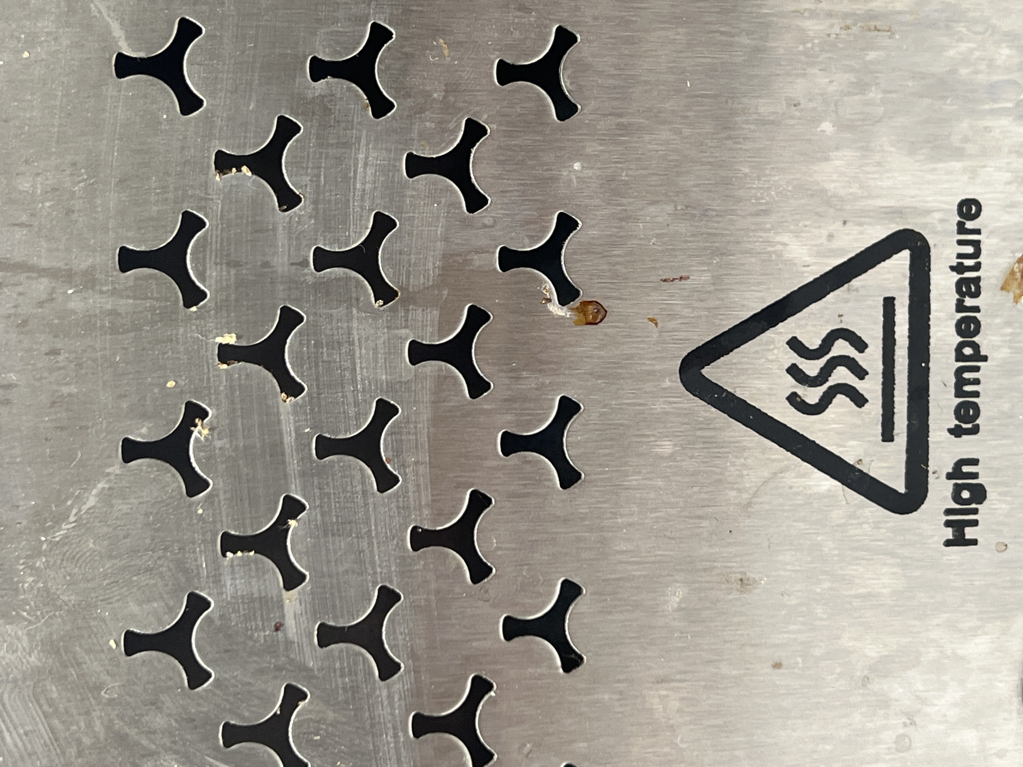 A close-up, vertically oriented image displays a metallic surface, likely brushed stainless steel, characterized by a repeating pattern of perforations and a warning label. The perforations are uniformly spaced, black, and shaped like three-pronged propellers or Y-shapes, cut through the metal.
The metallic surface exhibits signs of use, including faint scratches and smudges. Small, light-colored specks, resembling food crumbs or dried residue, are scattered across the surface and visible within some of the perforations. A more distinct, dark reddish-brown, somewhat viscous stain is prominent near the warning label.
On the right side of the frame, a black triangular warning symbol is present. Inside the triangle are three wavy, horizontal lines, universally recognized as a symbol for heat or steam. Below this symbol, the text "High temperature" is printed vertically in black capital letters.
The object is a static surface, possibly a component of an appliance or industrial equipment designed for heating or ventilation, such as a grill, oven grate, or food warmer, given the heat warning and perforations. No specific activity is taking place, nor are any people present. The debris suggests prior use. No discernible time of day, weather, or emotions are evident.
