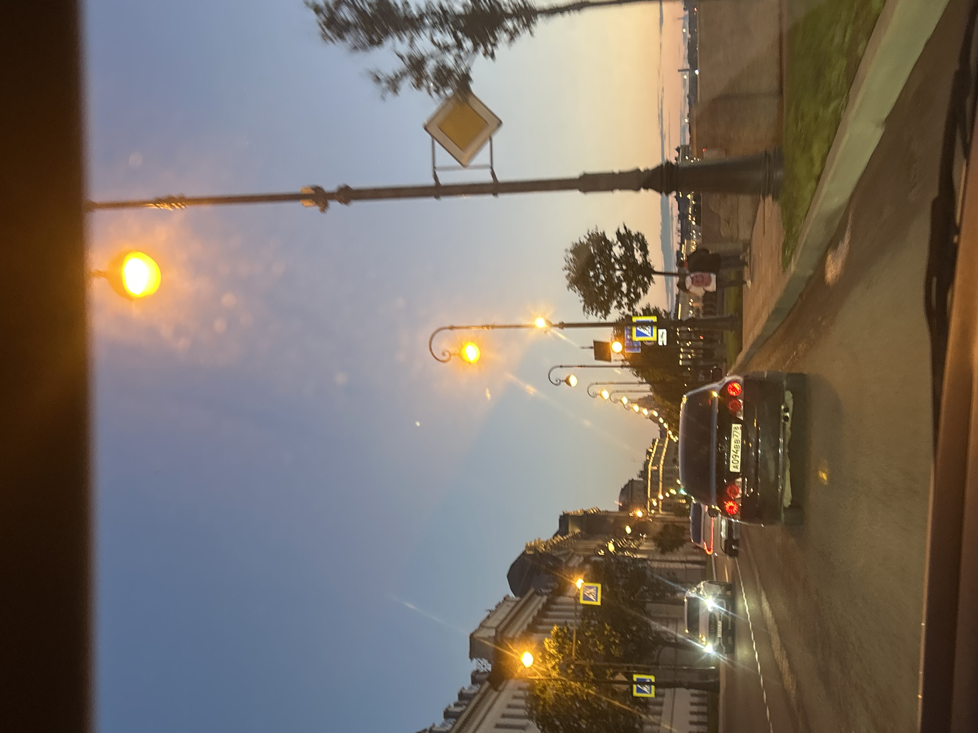 The image shows a street scene at dusk. Several cars are driving on the road, including a dark sedan in the foreground with its taillights illuminated. There are streetlights lining the road, some of which are lit with a warm yellow glow. Buildings with ornate architecture are visible on the left side of the street. In the distance, a body of water and buildings can be seen. Two pedestrians are walking on the sidewalk on the right side of the street. The sky is a gradient of blue and orange, indicating the time of sunset. There are traffic signs on poles along the road, including a diamond-shaped yield sign and pedestrian crossing signs. The overall impression is of a city street at the end of the day.