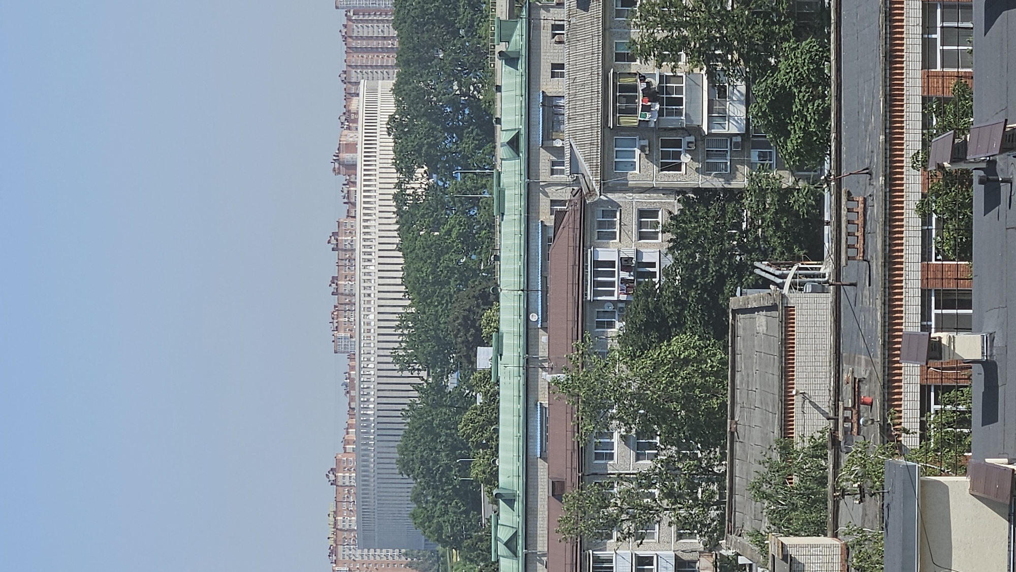 An aerial, long shot captures a sunny urban landscape on a clear day. The sky is a pale, cloudless blue. In the foreground, multiple apartment buildings are visible, varying in height and architectural style. The closest buildings have grey brick facades and dark, sloped roofs. Some of these buildings feature balconies with laundry hanging to dry. The middle ground is dominated by more apartment buildings with grey and light brown facades, topped with green and brown pitched roofs. Lush green trees are interspersed between and around these buildings. In the background, a large, modern, circular building with a white facade and numerous vertical elements stands out, surrounded by a line of taller, residential buildings with reddish-orange facades and white trim. The overall scene depicts a densely populated urban environment with a mix of older and newer constructions.