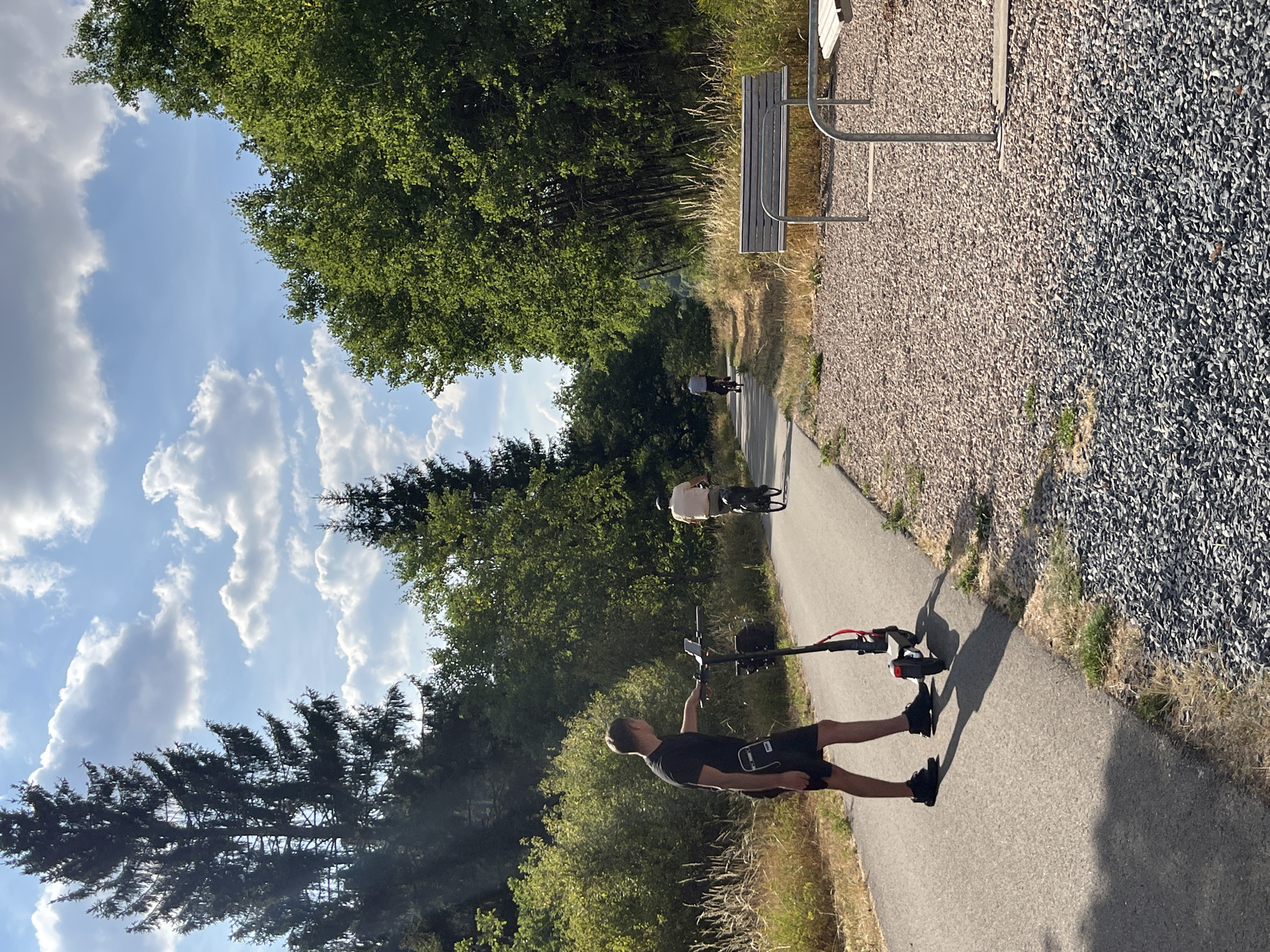 In Saint-Vith, Belgium, on a sunny day with a clear blue sky and scattered white clouds, a paved path winds through a wooded area. A man stands on the path, facing away from the camera and holding the handlebars of a red and black electric scooter. He is wearing a black t-shirt and black shorts. Further down the path, two individuals are cycling. The person in front is wearing a light-colored shirt and dark shorts, while the person behind them is wearing a light-colored shirt and dark pants. To the right of the path, a gravel area features a metal bench. Lush green trees and vegetation line both sides of the path, with tall evergreen trees visible in the background. The scene depicts a recreational pathway used for cycling and scootering, suggesting a park or a dedicated outdoor leisure space within the city.