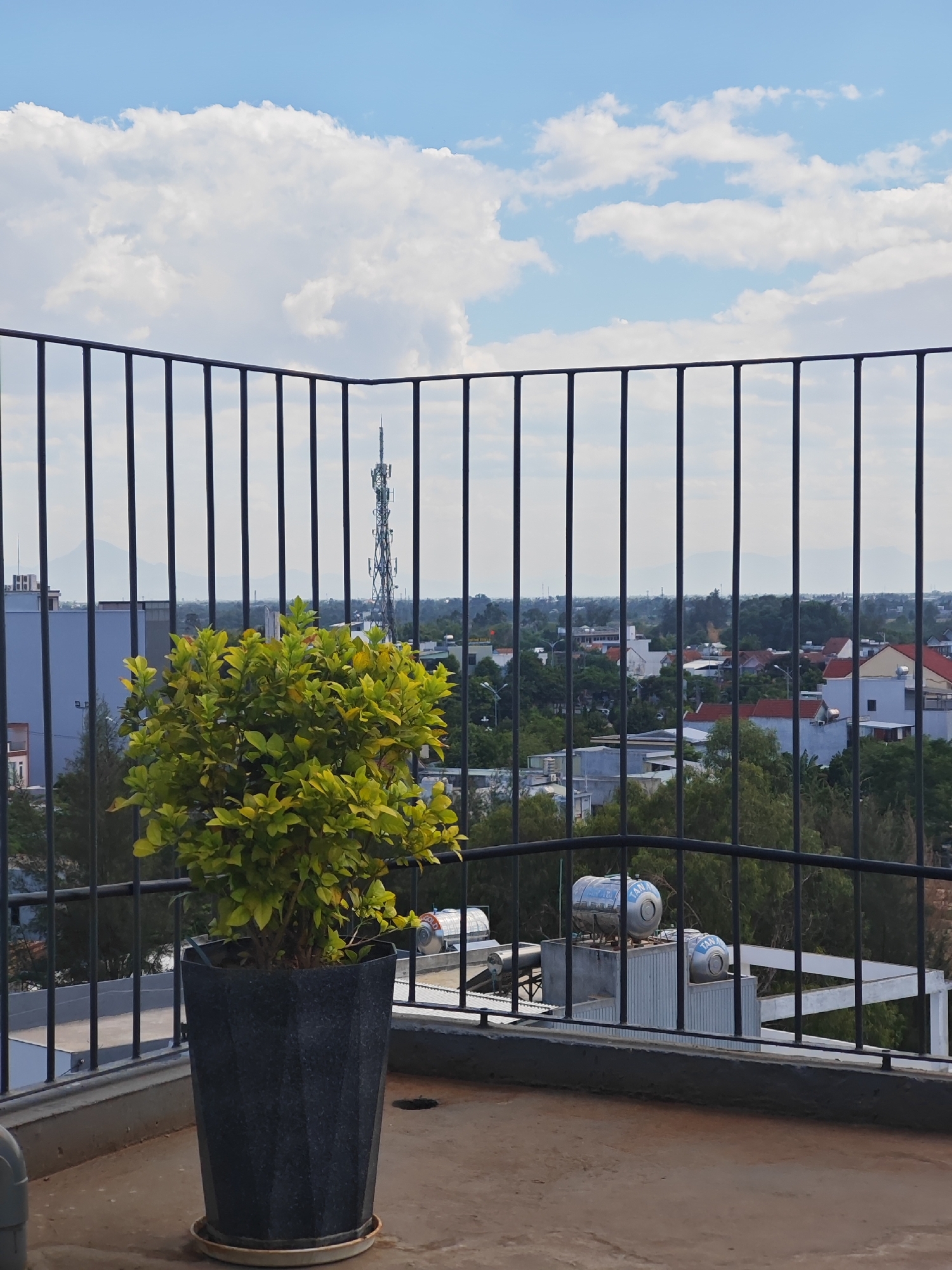 A view from a balcony in the city of Đà Nẵng, Vietnam, shows a potted green plant in the foreground. Behind the plant is a black metal railing. Beyond the railing, a cityscape unfolds with buildings, trees, and a distant transmission tower. The sky above is blue with scattered white clouds. Two large cylindrical tanks, likely water heaters, are visible on a nearby rooftop. The balcony floor is concrete.