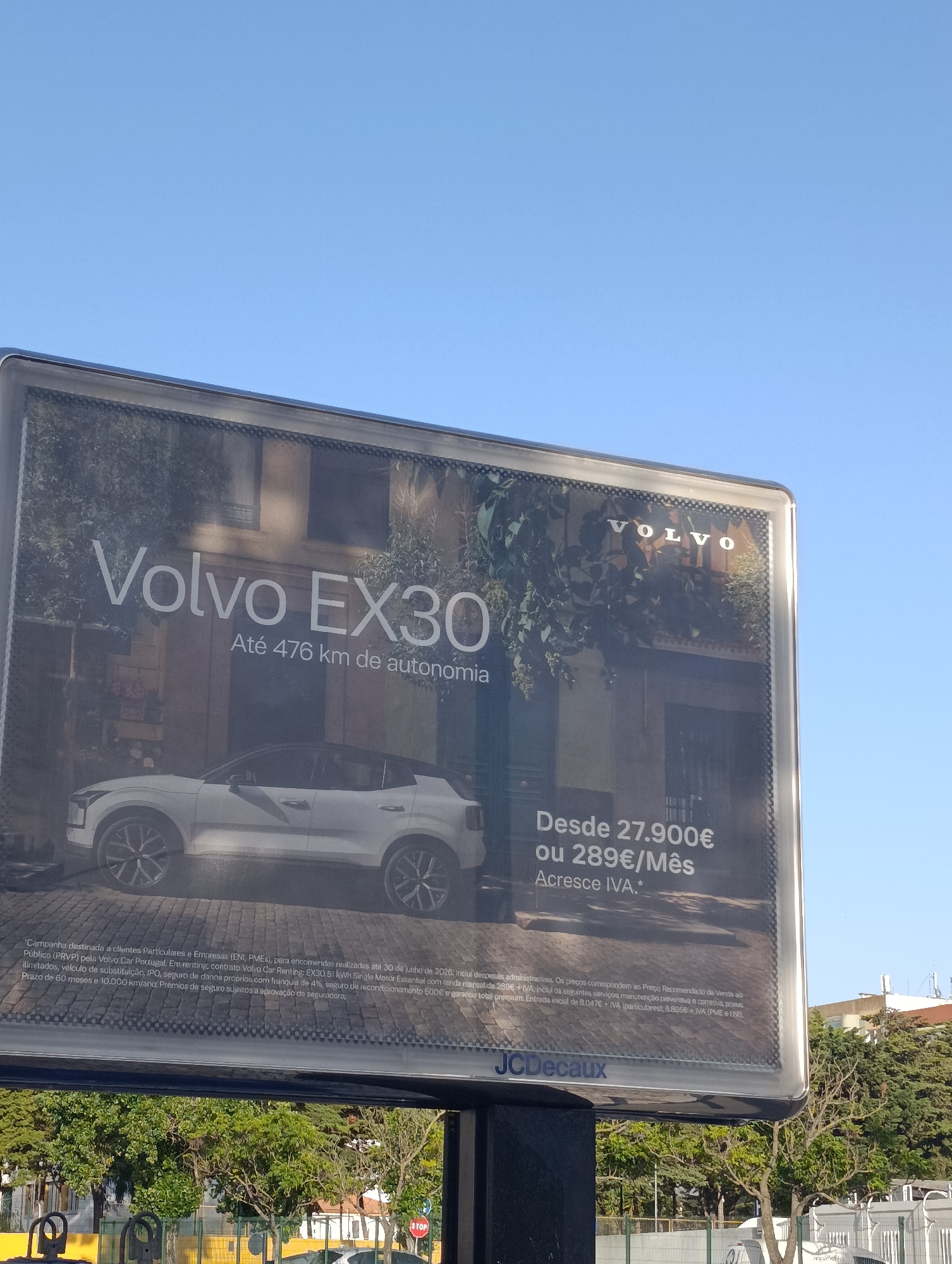 The image shows a large outdoor billboard advertising the Volvo EX30 car. The billboard features a white Volvo EX30 parked on a cobblestone street in front of a building with trees and foliage. The text on the billboard is in Portuguese and reads "Volvo EX30 Até 476 km de autonomia" (Volvo EX30 Up to 476 km of autonomy) and "Desde 27.900€ ou 289€/Mês Acresce IVA." (From €27,900 or €289/month Plus VAT.). The billboard also displays the Volvo logo and the JCDecaux logo at the bottom.
The billboard is mounted on a black pole and is situated outdoors, with a clear blue sky in the background. In the foreground, there are trees and some fencing, suggesting an urban or semi-urban environment. While the specific location in Setúbal, Portugal, is not explicitly stated, the presence of Portuguese text on the advertisement strongly indicates it is in Portugal. The scene appears to be a sunny day, with good visibility. There are no people clearly visible on the billboard itself or in the immediate vicinity, making it a static advertisement display.