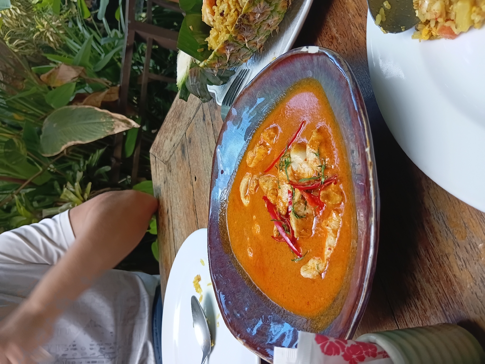 A close-up, overhead shot shows a meal laid out on a rustic wooden table at a restaurant in Baan Lamai, Thailand. The focus is on a bowl of vibrant orange curry filled with chunks of white fish, topped with thinly sliced red chilies and green herbs. To the right, a halved pineapple serves as a vessel for yellow fried rice, with a fork resting on a white plate beside it. In the foreground, a white napkin dispenser with a red floral pattern sits in a small green ceramic holder. A white plate with a spoon and scattered rice grains is visible to the left. In the background, out of focus, a person wearing a white t-shirt is seated at the table, their arms crossed. Lush green foliage and a glimpse of a railing suggest an outdoor seating area. The lighting is natural, casting a warm glow on the food and table.