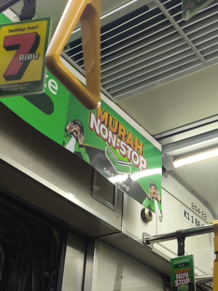 The image displays the interior of a KRL Commuterline train car, consistent with services operating to and from Cikarang, Indonesia. A yellow, inverted U-shaped handrail hangs from the ceiling in the upper left. To its left, a green and yellow sign with "Setiap hari 7 RIBU" (Every day 7 THOUSAND) and partially visible "GrabBike" branding is suspended. Across the upper wall, a prominent green and black banner advertises "GrabBike" with the red and white text "MURAH NON-STOP" (CHEAP NON-STOP) and "Cek sekarang!" (Check now!). This banner features a cartoon depiction of a man in a green helmet and jacket, gesturing with both hands, repeated twice. The off-white ceiling contains horizontal air vents and a linear fluorescent light fixture on the right. On the right wall, a white label displays "204-22" and the train car identification number "K1 1864". Below the main banner and to the right, a small, round, metallic object, possibly a speaker, is affixed to the wall. Further right, a small green "GrabBike MURAH NON-STOP" sign hangs from a horizontal metal rod. A metallic door frame is partially visible on the far left.