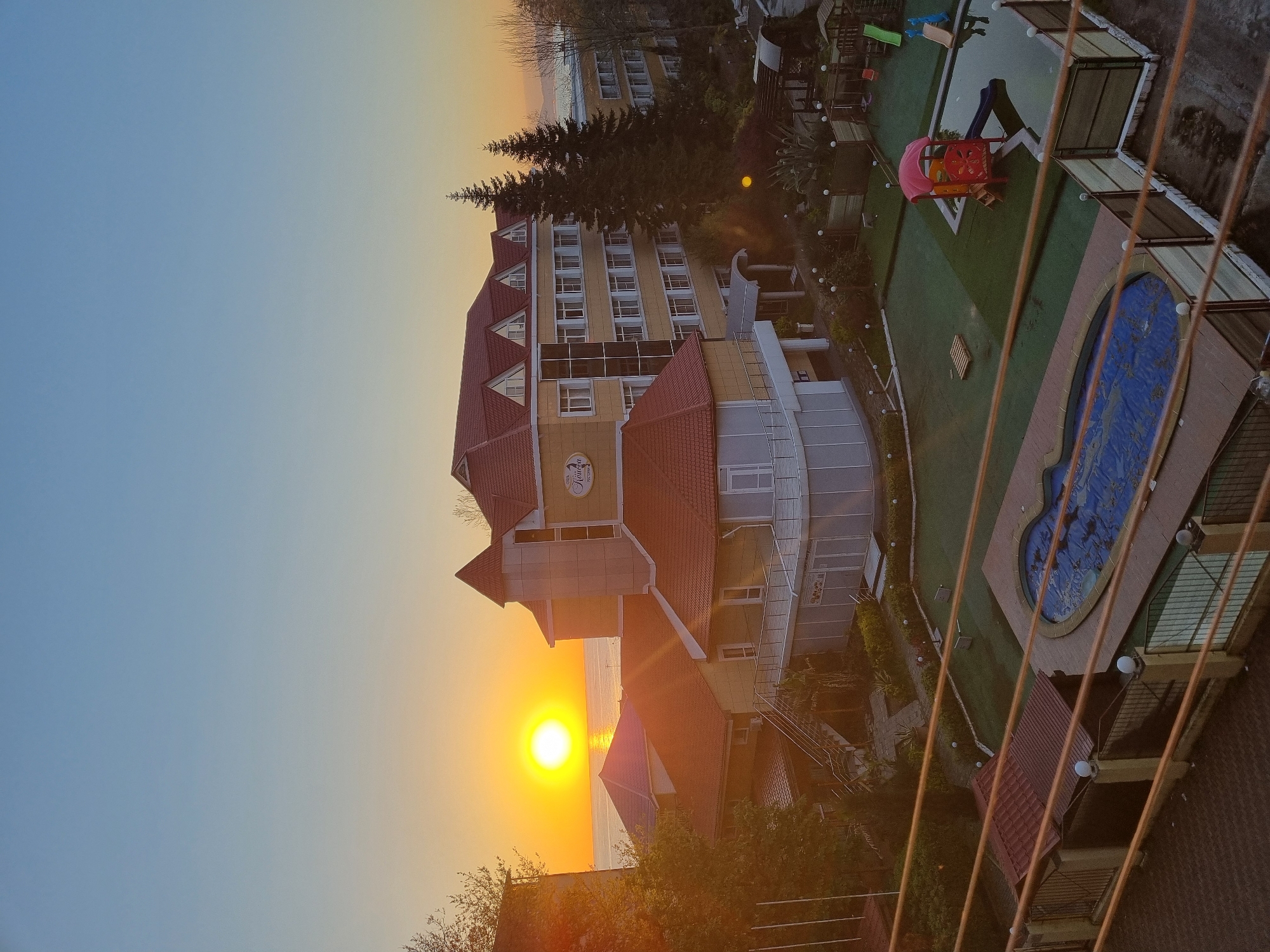 The image presents a tranquil aerial view of a coastal resort or hotel property, captured during either sunrise or sunset. The sky displays a beautiful gradient, transitioning from a clear light blue on the right to a vibrant orange and yellow on the left, where a brilliant, circular sun glows intensely near the horizon. This golden light reflects brightly on the calm surface of a large body of water, likely the Black Sea, stretching into the distance.
In the center-right of the frame, a multi-story hotel building with distinctive red-tiled roofs and light yellow walls is prominent. A sign on the building's facade clearly shows the Cyrillic text "ОТЕЛЬ "КОРОНА"" (HOTEL "KORONA"). Two tall evergreen trees stand near the hotel, and numerous windows dot its exterior.
Below and to the right of the main building, the hotel grounds feature a well-manicured green lawn. A children's playground with a colorful red and pink structure, including a slide, and a separate blue slide, is visible on the grass. Adjacent to the playground, a swimming pool with intricate blue mosaic tiling appears to be covered or partially filled. Paved pathways, outdoor lighting, and landscaped bushes complete the serene scene. The perspective is from an elevated position, possibly a balcony, as indicated by the diagonal lines (likely railings or wires) in the immediate foreground. The overall setting suggests a peaceful leisure destination in Sochi, Russia, bathed in the warm glow of the golden hour.