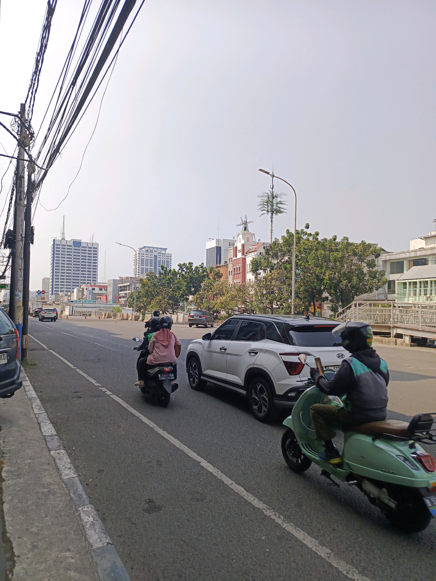The image captures a street scene in West Jakarta, Indonesia, under a hazy sky. The foreground is dominated by a light green Vespa scooter carrying a person wearing a dark jacket with teal accents and a helmet. They are riding on the right side of a multi-lane road. To the left of the scooter, a white Hyundai Creta SUV is also in motion. Further down the road, another scooter with two riders, one in a pink headscarf, is visible. The road is lined with a sidewalk and a gray curb. In the background, several buildings are visible, including tall office buildings and some with more distinct architectural features, such as a red and white building with a tower. Utility poles with numerous electrical wires are prominent on the left side of the frame. The general atmosphere suggests a typical urban environment with ongoing traffic.